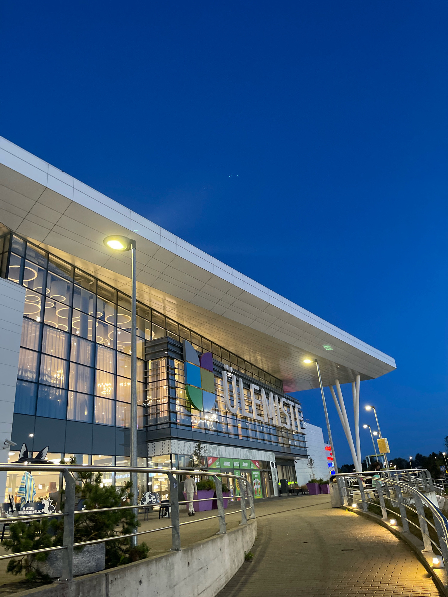 The image captures the Ülemiste Keskus, a shopping mall located in Tallinn, Estonia. The scene is set at dusk or early evening, with a deep blue sky overhead. The mall features a modern architectural design with a large glass facade, illuminated from within, showcasing decorative circular light fixtures. A prominent sign spelling out "ÜLEMISTE" in large, illuminated letters is visible on the building's exterior.
In the foreground, a paved walkway curves towards the mall entrance. A metal railing borders the walkway, with small lights embedded along its base, casting a warm glow on the ground. Several tall streetlights stand in front of the mall.
Outside the mall, some trees and large purple planters are placed. A few people are visible in the distance, walking along the pathway or near the mall entrances. One person is walking away from the camera on the right side of the pathway, while others are seated at tables outside what appears to be a cafe or seating area. The overall atmosphere is calm and urban, indicating a commercial area.