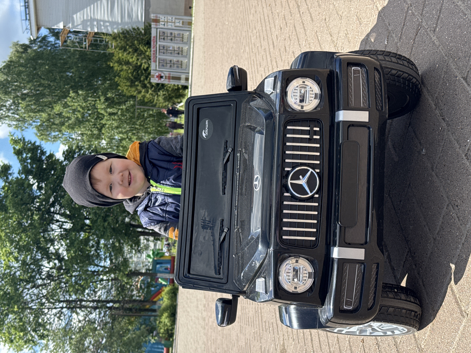 A young child in a black knit hat and a blue and yellow jacket smiles from behind the "Teknum" branded door of a black Mercedes G-Wagon toy car. The car is parked on a brick surface, with a building, trees, and playground visible in the background. Based on the presence of a "АЛЛЕЯ ПОМЯТИ" (Alley of Memory) sign in the background, this photo was likely taken in Zav'yalovo, Russia.