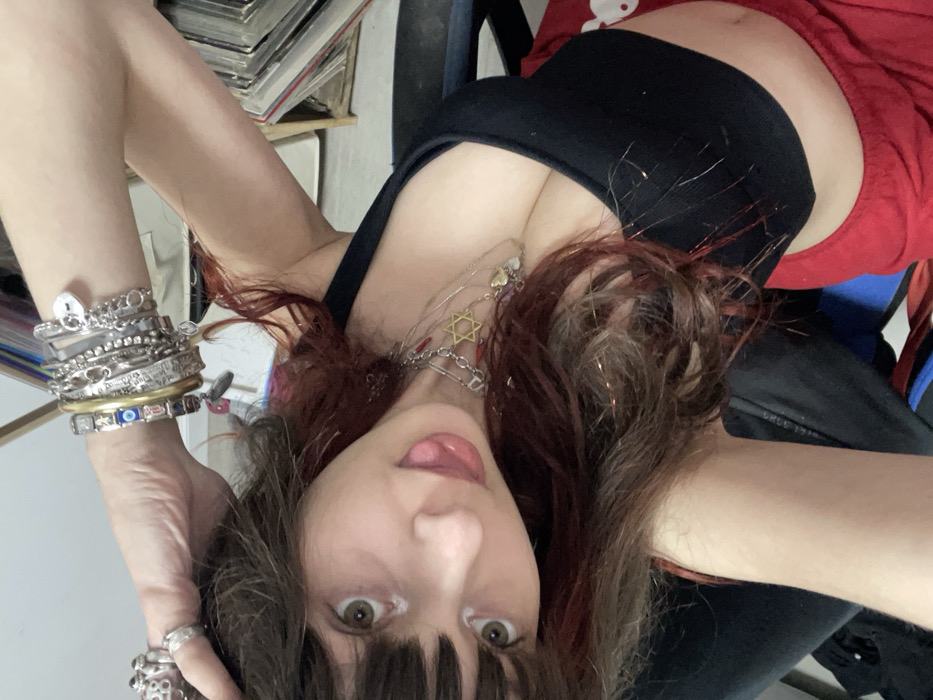 A woman with brown and red hair, wearing a black tank top and red shorts, is angled upside down. She's sticking her tongue out slightly. Her hair has glittery strands. She wears multiple silver bracelets on her left wrist and rings on her fingers. A Star of David necklace is visible. Books and a chair are in the background.