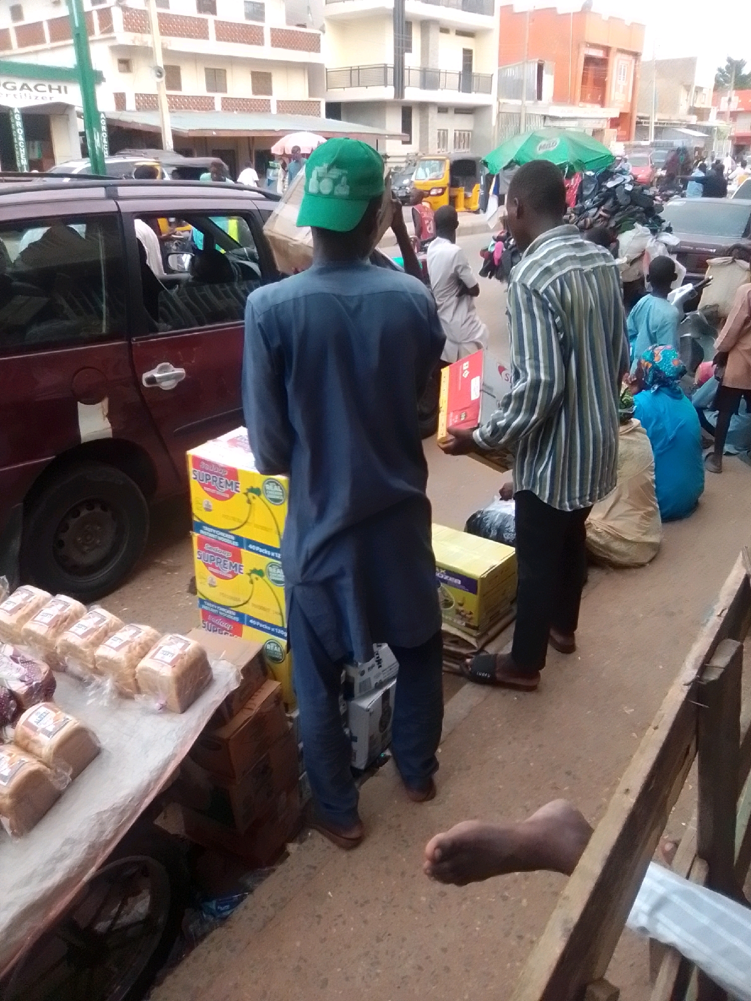 The image depicts a street scene in Arawa, Nigeria. In the foreground, a person wearing a blue shirt and pants and a green cap stands near a maroon vehicle. They are positioned to the right of a display of bread loaves and several yellow boxes stacked on top of each other. To the right of the first person, another individual in a striped shirt and black pants holds a box. Further to the right, a person in a blue garment sits next to a pile of items. In the background, buildings and various vehicles, including a yellow auto-rickshaw, are visible. The ground is paved with a rough surface.
