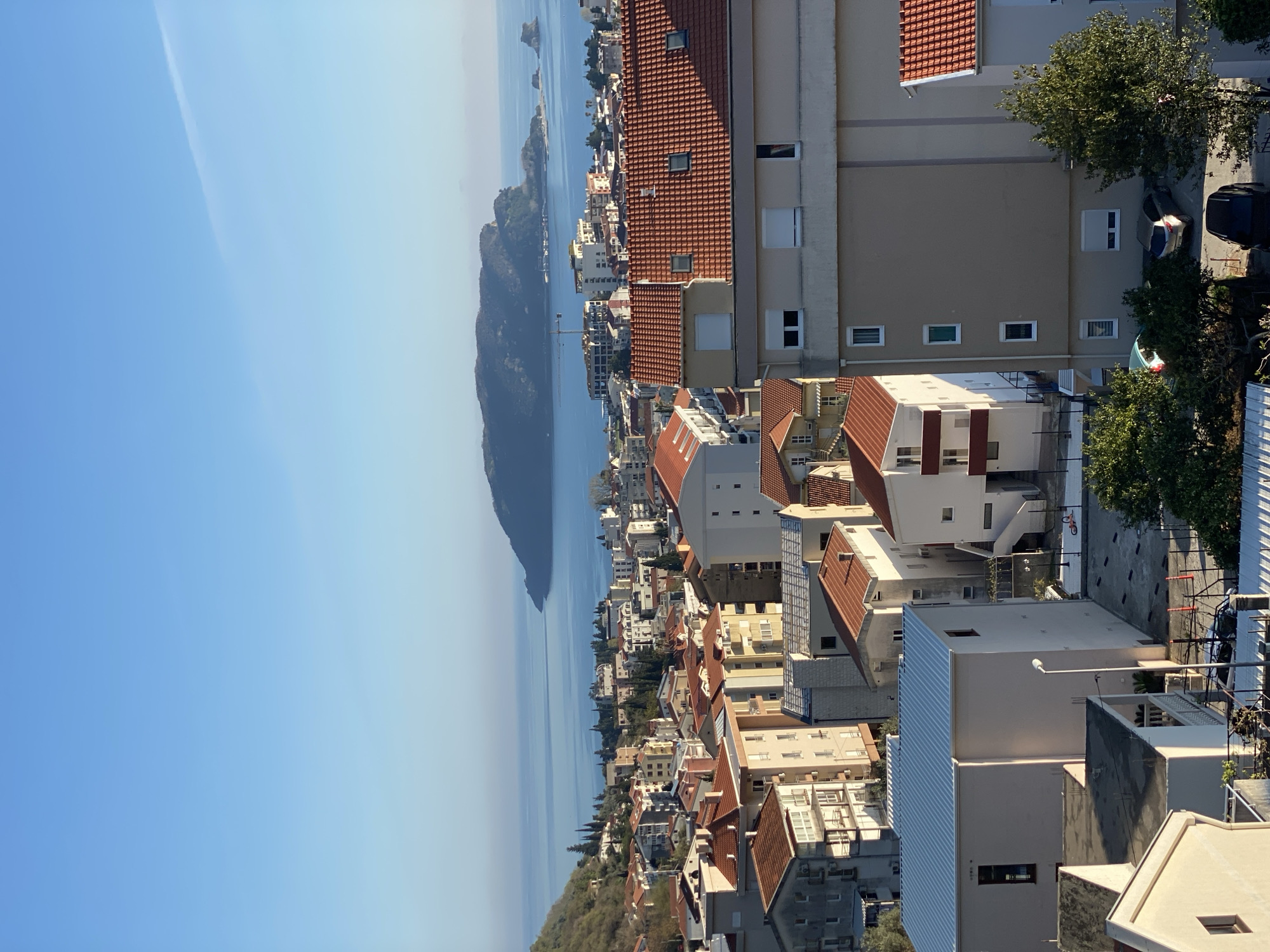 The image depicts a coastal view of the city of Budva, Montenegro. The scene includes a body of water, possibly the Adriatic Sea, with an island visible in the distance. The cityscape consists of buildings with terracotta roofs, interspersed with other structures. In the foreground, there are several modern buildings. The sky is clear blue. There are no visible people or interactions.