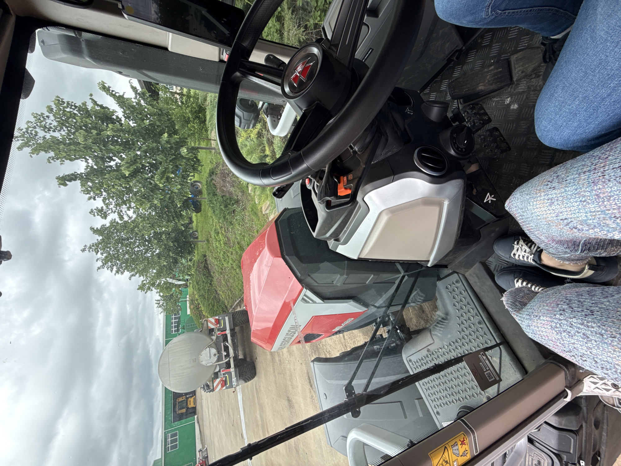 A view from inside the cab of a Massey Ferguson 8S.425 tractor, showing the interior elements: steering wheel with the Massey Ferguson logo, dashboard, floor pedals, and seat. Outside the cab, a green building is visible on the left, along with a large water or liquid tank trailer. The background includes green grass, trees, and a cloudy sky. The photo was likely taken in an agricultural or industrial setting. The location of Gorna Mitropolia, Bulgaria is implied.