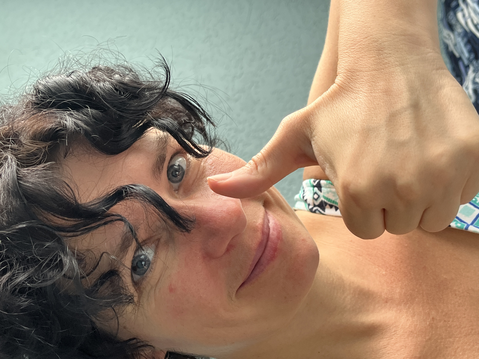 A close-up, waist-up shot shows a person with curly dark hair and blue eyes giving a thumbs-up. They are looking directly at the camera and smiling slightly. The person is wearing a dress with a geometric pattern. The background is a muted teal wall. The image appears to have been taken indoors. There is no information that can be used to identify the location in the city of ღრმაღელის თემი, Georgia.