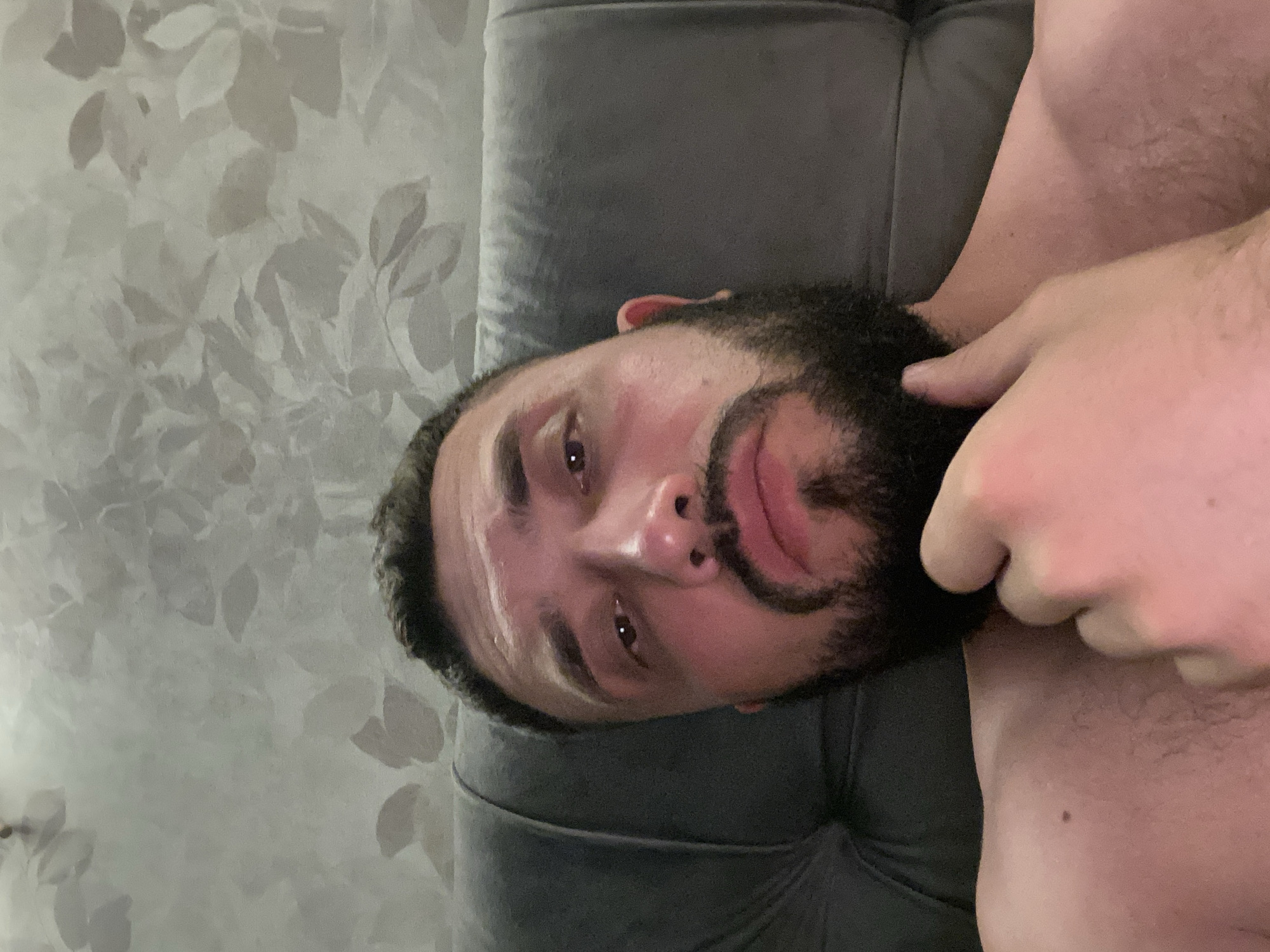 The image is a close-up, selfie-style photograph of a man with a beard, lying down. He is positioned against a dark gray, tufted headboard, with a textured wallpaper featuring a floral pattern visible behind him. The man's right hand is resting on his chin and jawline, and he is looking directly at the camera with his eyes open. His expression is neutral. The lighting appears to be artificial and casts a pinkish hue on his skin. The lower portion of the image shows his bare chest and shoulder. There is no indication of the specific location in Derbent, Russia, nor any context suggesting it is a crime scene.