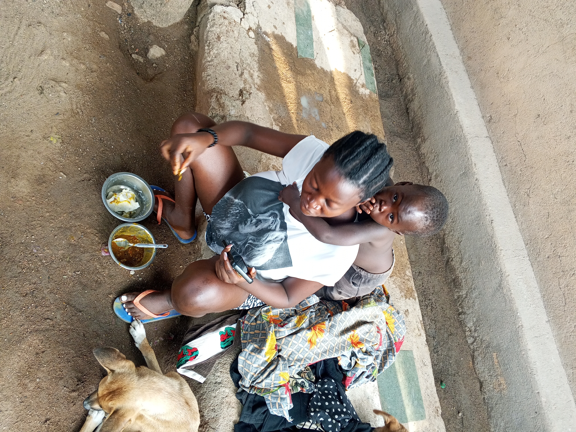 A top-down medium shot shows a woman sitting on a concrete step. A child clings to her back. The woman is holding a phone, and a dog is lying nearby. Two metal bowls containing food are visible beside her. The location is Jos, Nigeria.