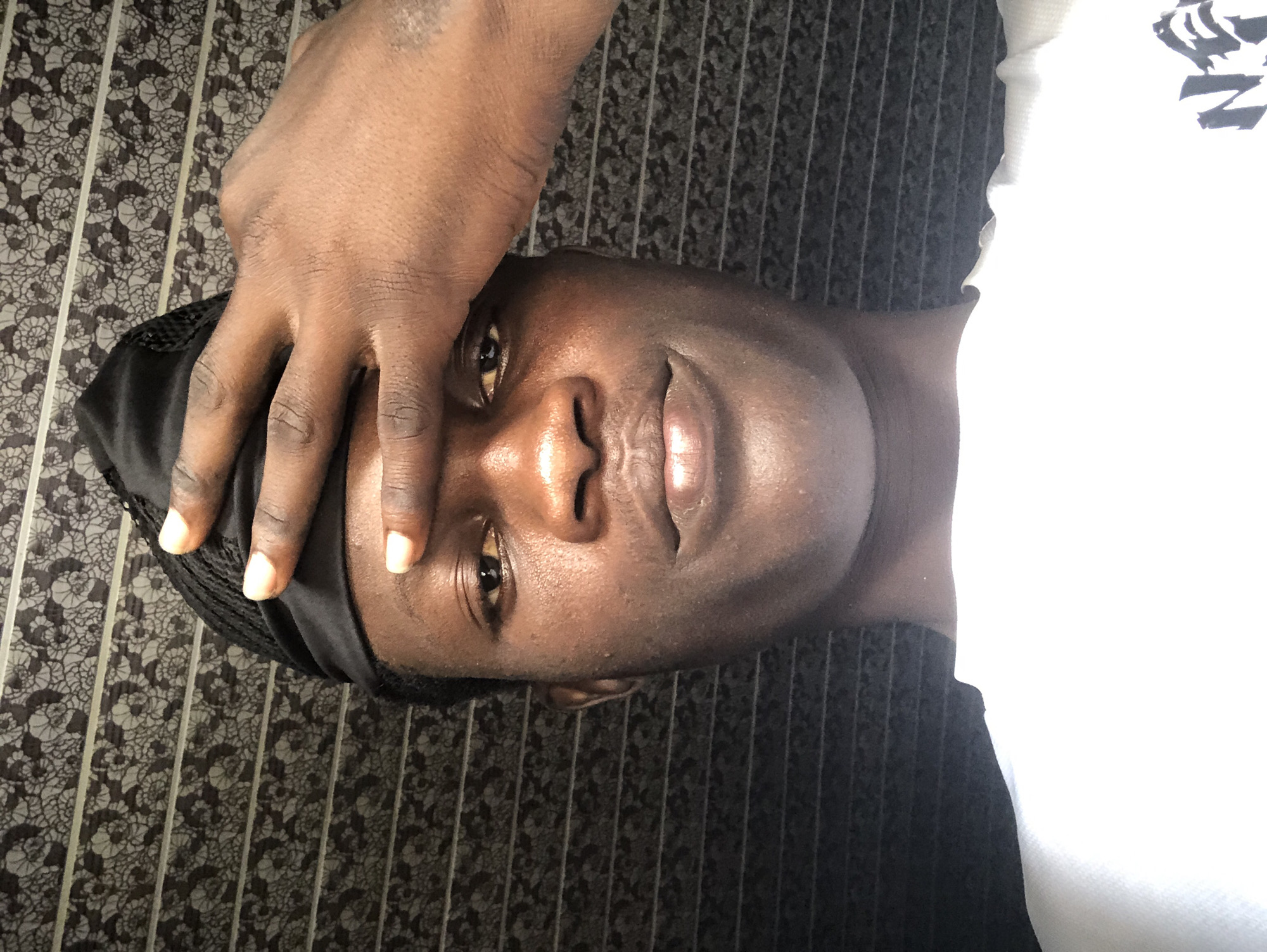The image is a close-up, selfie-style shot of a dark-skinned individual with a black durag on their head. Their right hand is covering their forehead. The background consists of a patterned wallpaper or fabric with a repeating floral design. The individual is wearing a white t-shirt with a black logo on the right side. The lighting suggests an indoor setting. The image does not provide any specific location in Ado, Nigeria, or any contextual details beyond the visual elements present.