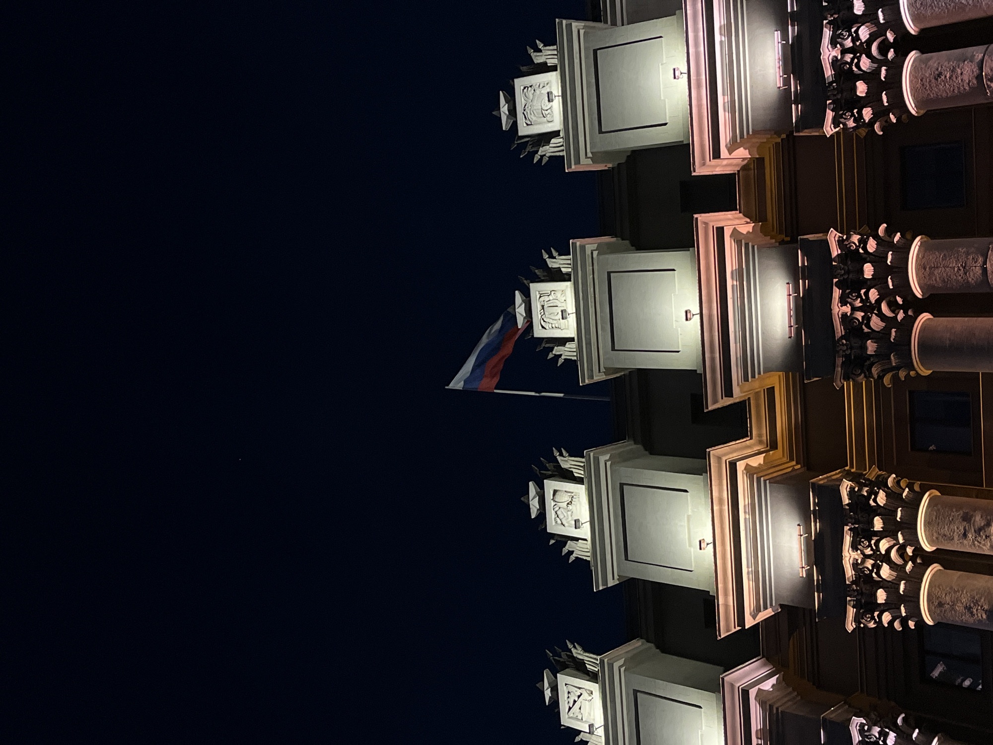 The image shows the illuminated facade of a building at night in Novokuybyshevsk, Russia. The upper portion of the building features decorative elements, including what appear to be stylized stars and possibly coat of arms carvings, which are highlighted by spotlights. A Russian flag is visible flying from a flagpole atop the structure. The lower portion of the building is characterized by ornate columns with intricate carvings, also illuminated by warm-toned lights. The sky above is dark, indicating nighttime. There are no people or any discernible actions or interactions visible in the image.