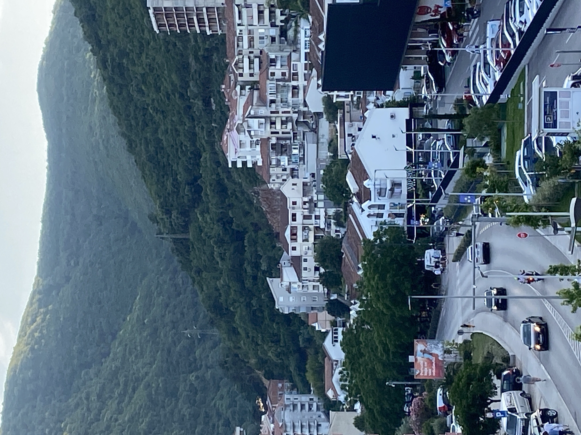 An aerial or elevated perspective captures a bustling town nestled at the base of a verdant, densely forested mountain range, likely in Budva, Montenegro, during what appears to be late afternoon or early evening. The mountain slopes, covered in dark green foliage, recede into the distance under a bright, overcast sky.
In the mid-ground, a dense cluster of multi-story buildings, predominantly white or light-colored with red or orange tiled roofs, extends along the mountain's contours. These structures display a mix of modern and traditional architecture, with numerous windows and balconies. Trees and other vegetation are interspersed among the buildings, adding greenery to the urban landscape.
The foreground features a wide, curving asphalt road with white markings, indicating traffic lanes and crosswalks. Numerous vehicles, including cars and vans, are visible both parked along the road and in motion. A motorcyclist is also seen on the road. On the right side, there's a parking area with multiple cars. Street lights and utility poles line the thoroughfare.
Visible details include a red hexagonal "STOP" sign on a pole beside the road. A large outdoor billboard displays an image of a person performing a water sport, likely wakeboarding or surfing, with partially legible text below, including words that appear to be in a South Slavic language (e.g., "naj", "Podgorica" or similar is hinted). Other smaller signs, such as one with a "P" for parking and what looks like "VI PISA" on a building, are present. A small open green space with scattered trees and bushes is also visible near the road, along with various signs and banners for businesses or services. Several pedestrians are sparsely seen on sidewalks or near parked vehicles, contributing to the sense of an active urban environment.