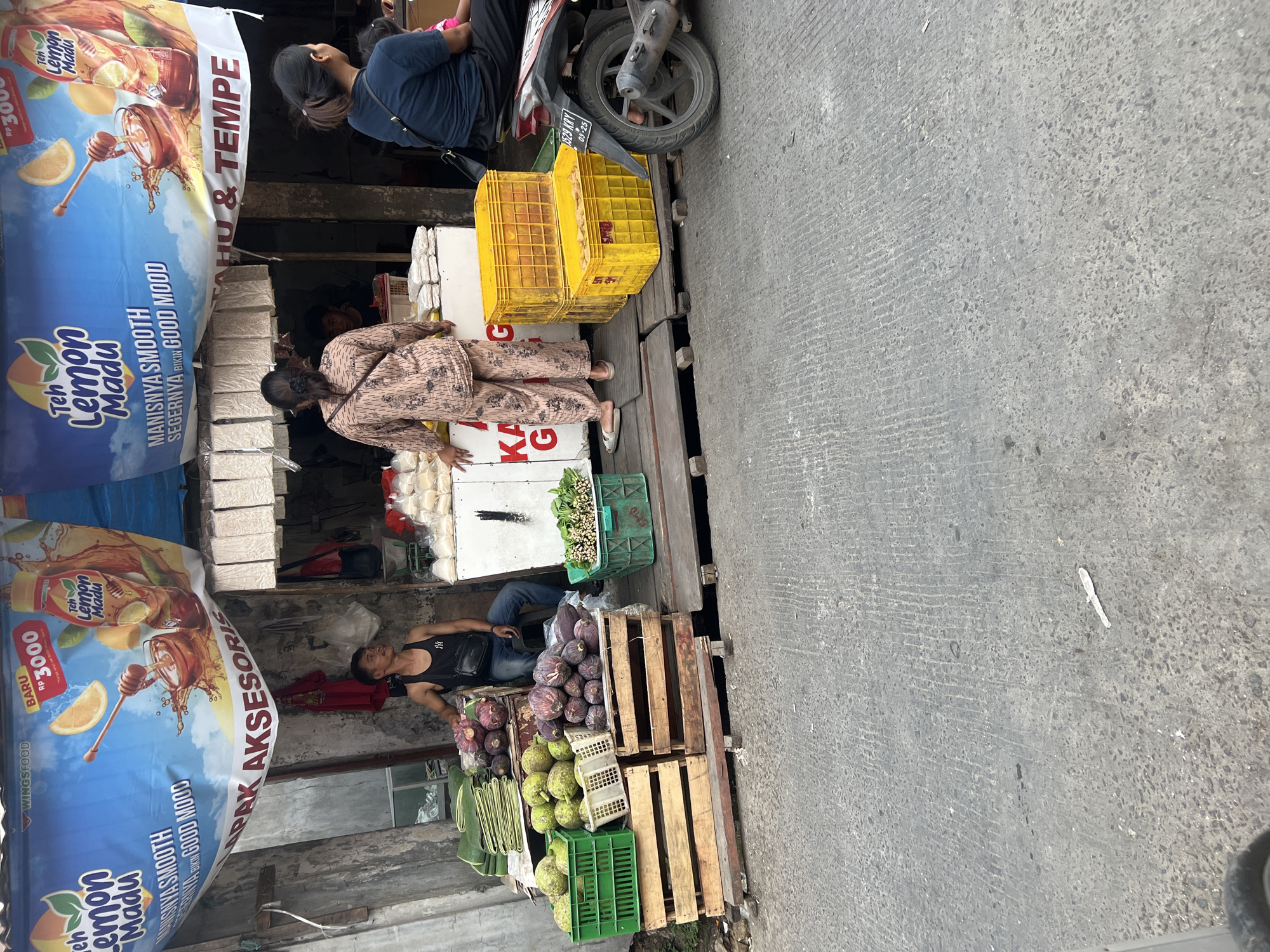 A street vendor stall in Citeureup, Indonesia, displays a variety of produce and packaged goods. In the foreground, a man in a black tank top and jeans sits to the right of a display of green bananas and reddish-purple fruits, possibly bananas or plantains. To his left are wooden crates holding more of these fruits and large green produce. Further to the left, a green plastic crate contains leafy green vegetables. Behind the man, a woman in a patterned outfit stands near a stall with packaged goods and yellow crates stacked on top of each other. The stall has banners advertising "Teh Lemon Madu" (Lemon Honey Tea). A motorcycle is parked to the right of the stall. The ground is paved with a textured gray asphalt. The scene is outdoors with a weathered building in the background.