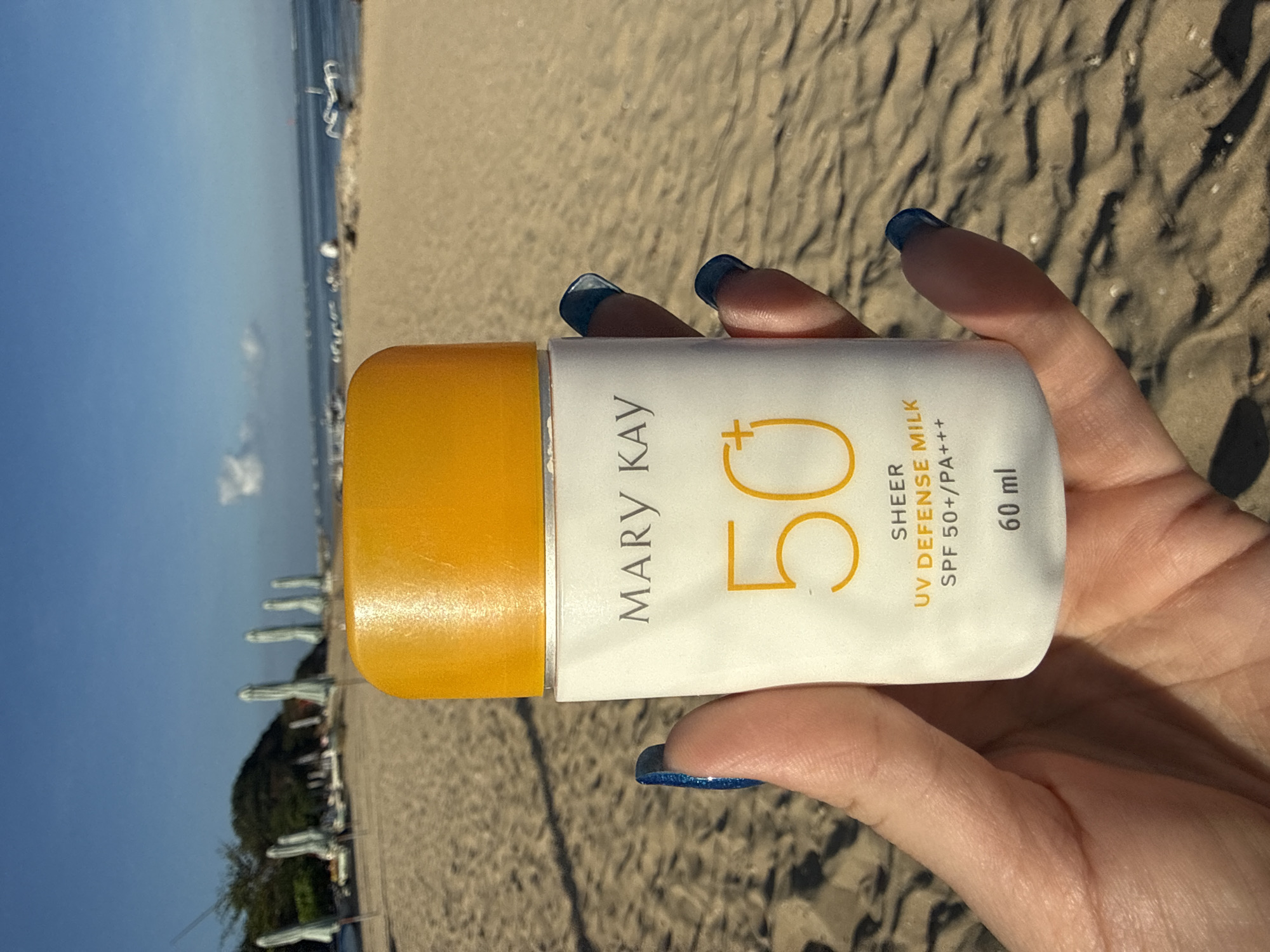A close-up shot shows a hand holding a white bottle of Mary Kay UV Defense Milk SPF 50+. The bottle has an orange cap and the text "MARY KAY", "50+", "SHEER UV DEFENSE MILK SPF 50+/PA+++", and "60 ml" are visible. The background is a sandy beach with a calm ocean and clear blue sky. Several beach umbrellas and loungers are visible in the distance. The setting appears to be a beach in Denpasar, Indonesia, a popular tourist destination known for its beautiful coastline.
