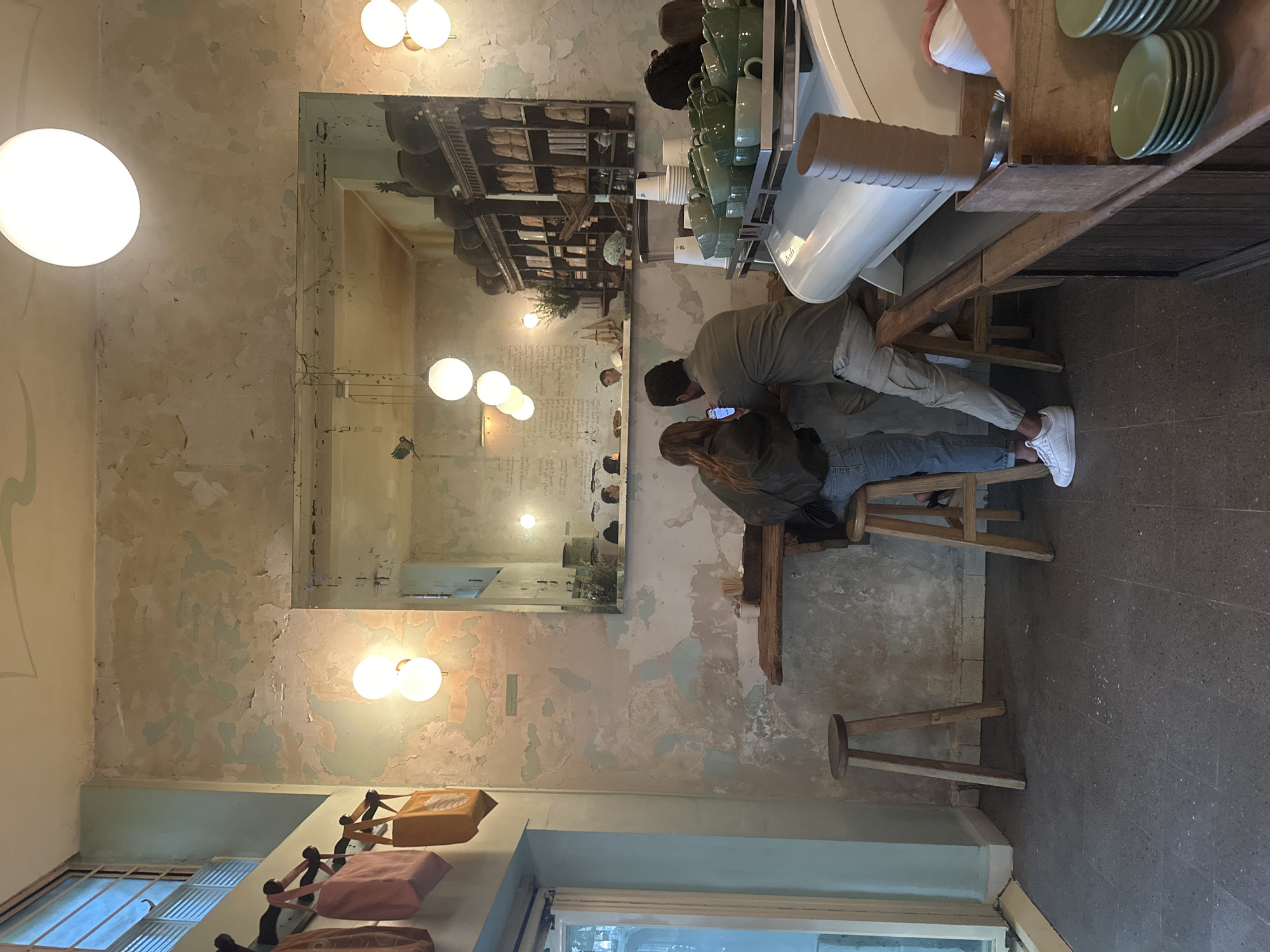 The image shows the interior of a cafe in Mexico City, Mexico. The walls are textured and have a distressed appearance with patches of faded paint. On one wall, there is a large mirror reflecting a scene of shelves filled with books. Light fixtures with round bulbs are mounted on the walls.
Two people are present in the image, standing by a counter. One person is looking at a mobile phone while the other is standing next to them. A counter is topped with a beverage machine, cups, and plates. Wooden stools are placed on the floor. Near the bottom of the image, there is a window and shopping bags.