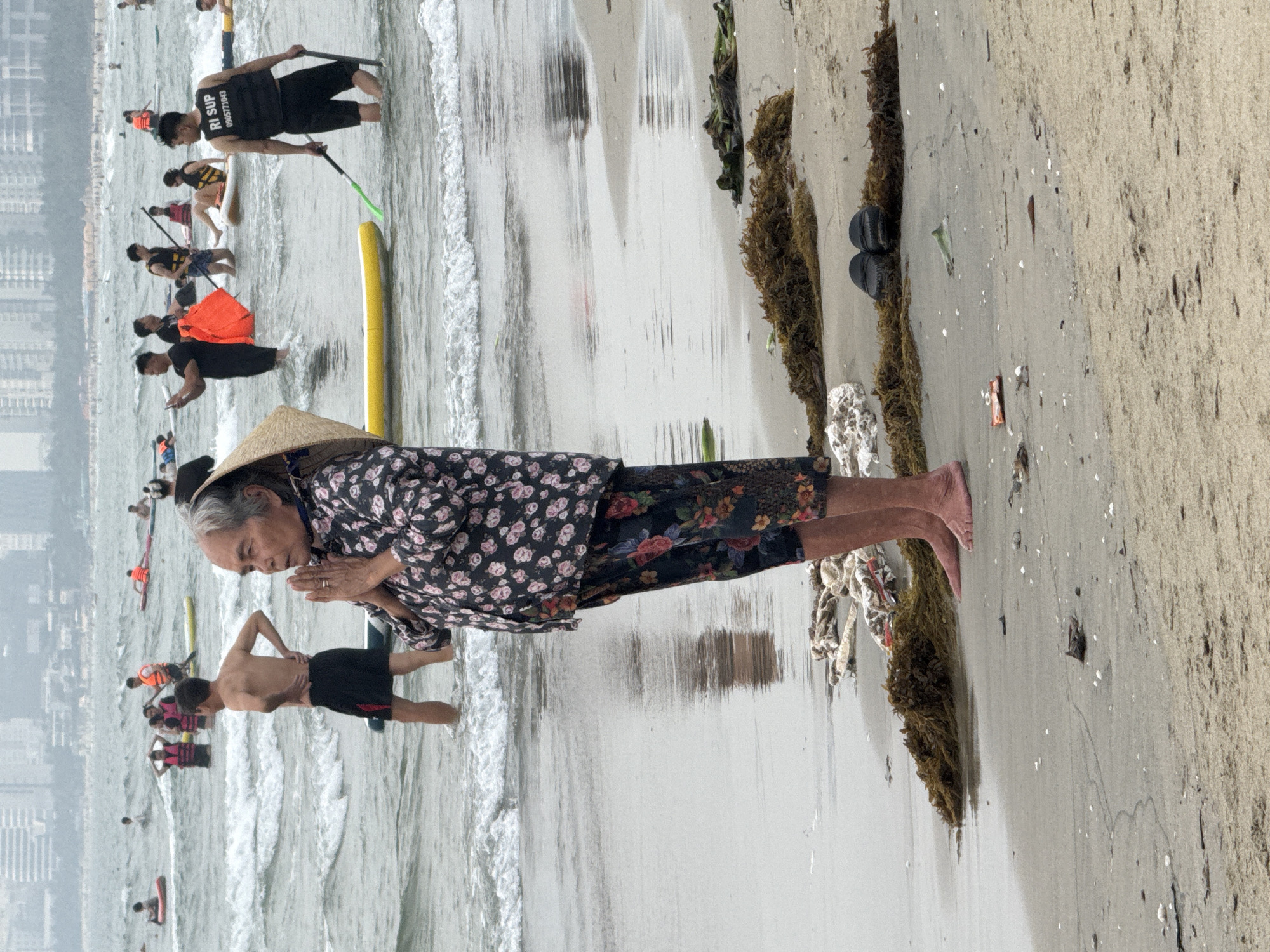 The image depicts a beach scene in Đà Nẵng, Vietnam. An elderly woman wearing a conical Asian hat, a black floral shirt, and dark floral pants stands with her hands clasped in a prayer-like gesture. Her bare feet rest on the wet sand.
Behind her, the beach extends with seaweed and debris scattered along the shoreline. Further back, the ocean is dotted with people participating in water activities, including paddleboarding. Several of these individuals wear life vests. In the distance, buildings line the coastline, defining the urban backdrop of Đà Nẵng.