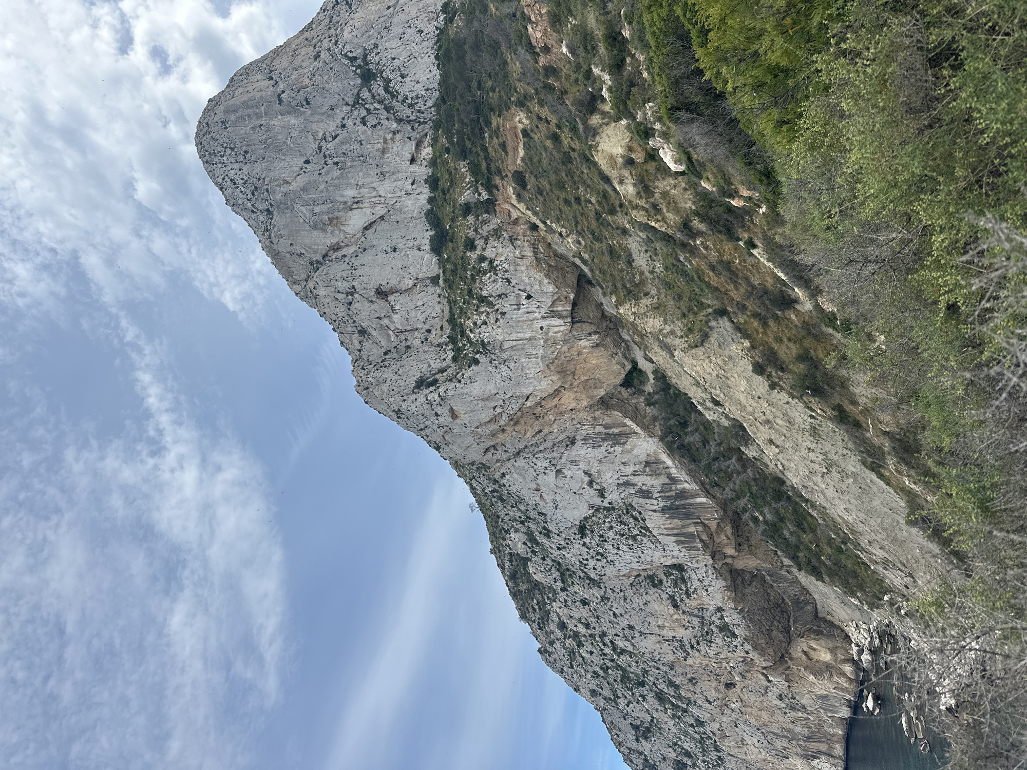 The image features the Penyal d'Ifac, a massive limestone rock formation, located in Calp, Spain. The rock face is characterized by its steep, rugged terrain with varying textures and colors ranging from light gray to beige. Patches of green vegetation cling to the rock face, especially along the right edge of the frame. The sky, visible on the left side of the image, is pale blue with scattered white clouds. At the base of the rock, there is a glimpse of dark water.
