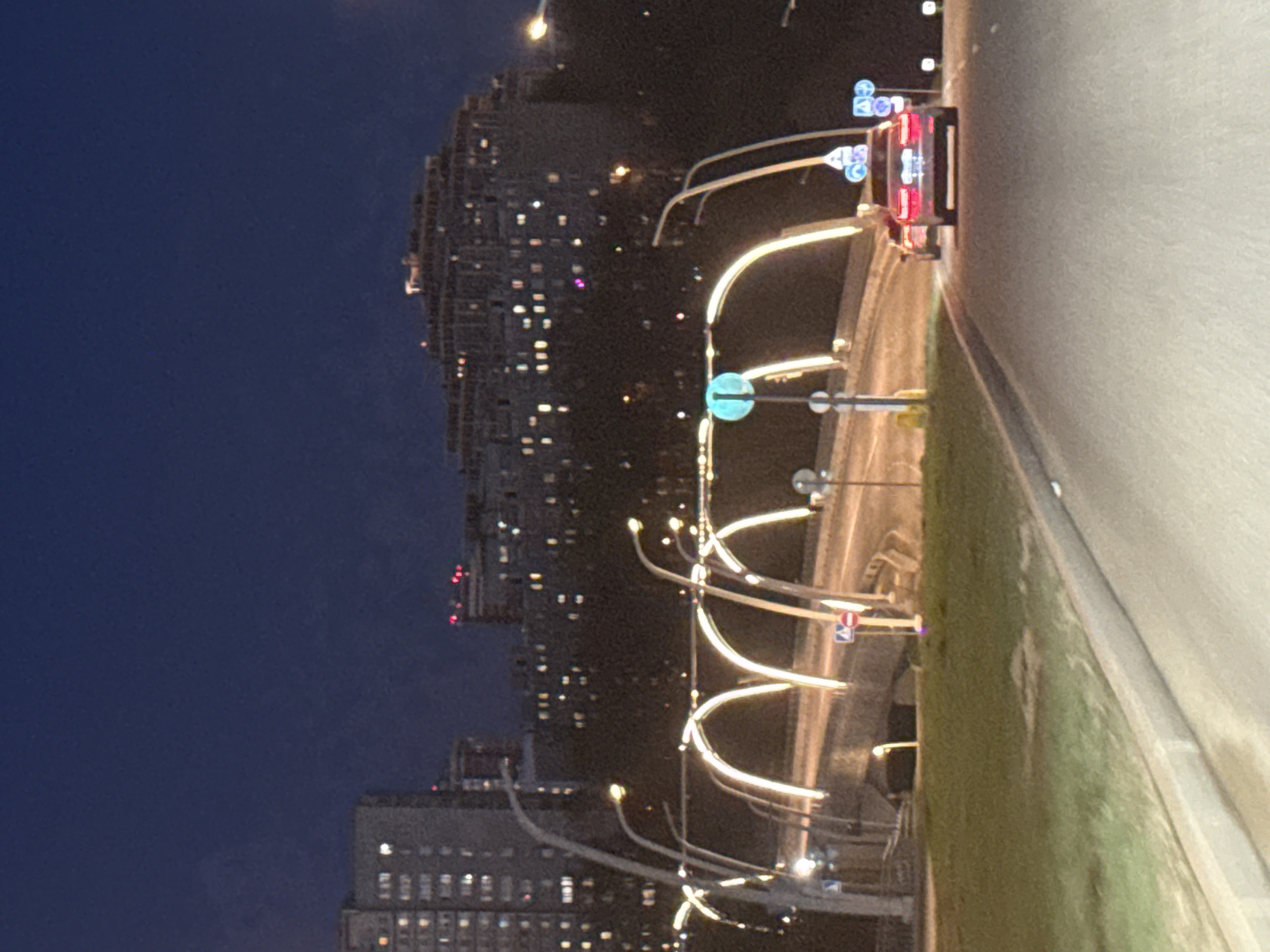 The image captures a nighttime urban or suburban scene in Khimki, Russia. On the left, a cluster of modern, multi-story residential buildings rises against a dark blue night sky. Numerous windows in these buildings are lit, indicating occupancy and creating a pattern of bright dots.
To the right, a multi-lane highway or main road stretches into the distance, illuminated by a series of distinctive streetlights. These lights feature prominent arched supports and emit a bright, warm white glow, casting light onto the road surface and a grassy median strip. On the road, a vehicle, likely a car or van, is visible with its bright red taillights illuminated, suggesting movement or a stopped position. Other faint lights in the distance indicate further traffic.
Along the roadside and above the highway, several traffic signs are visible. These include circular blue signs, typically denoting mandatory instructions, and rectangular red and white signs, which are often regulatory or informational. Due to the distance and image resolution, the specific text or symbols on these signs cannot be deciphered. The overall atmosphere is calm, with the artificial lights creating a clear and well-lit nocturnal environment. The time of day is clearly night, with no visible adverse weather conditions.