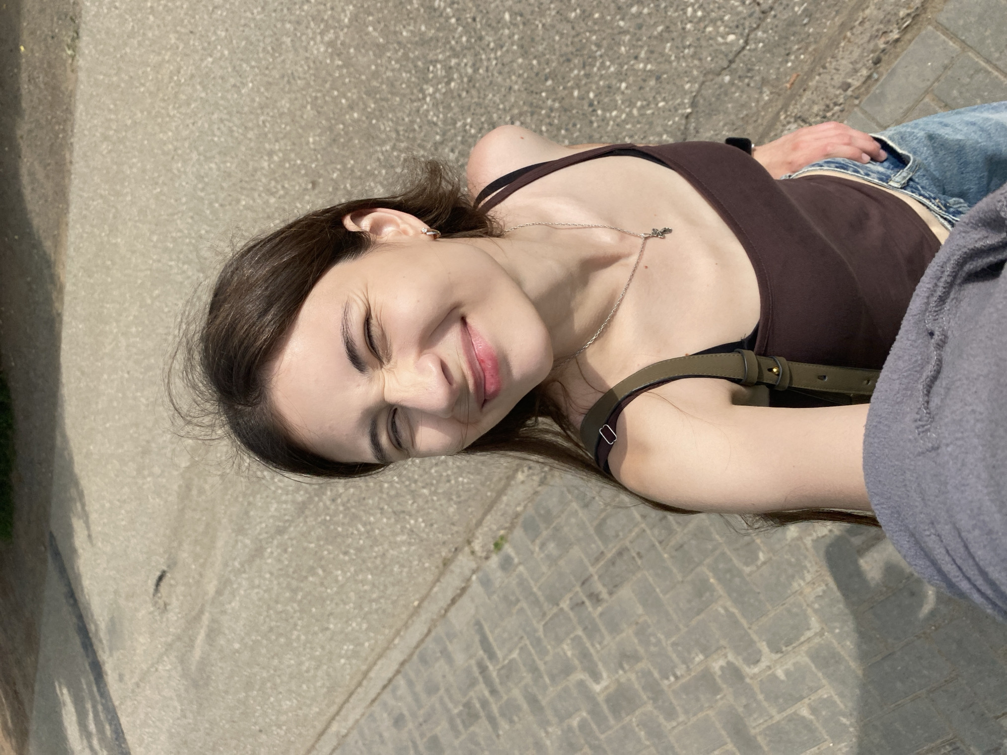 An overhead shot shows a young woman with brown hair and light skin, squinting and smiling as she holds her phone to take a selfie. She is wearing a brown tank top, blue jeans, and a silver necklace with a cross pendant. A green strap from a bag is visible over her right shoulder. The background consists of a paved sidewalk on the left and a textured asphalt surface on the right. The lighting suggests it is daytime. The scene appears to be an outdoor public space.