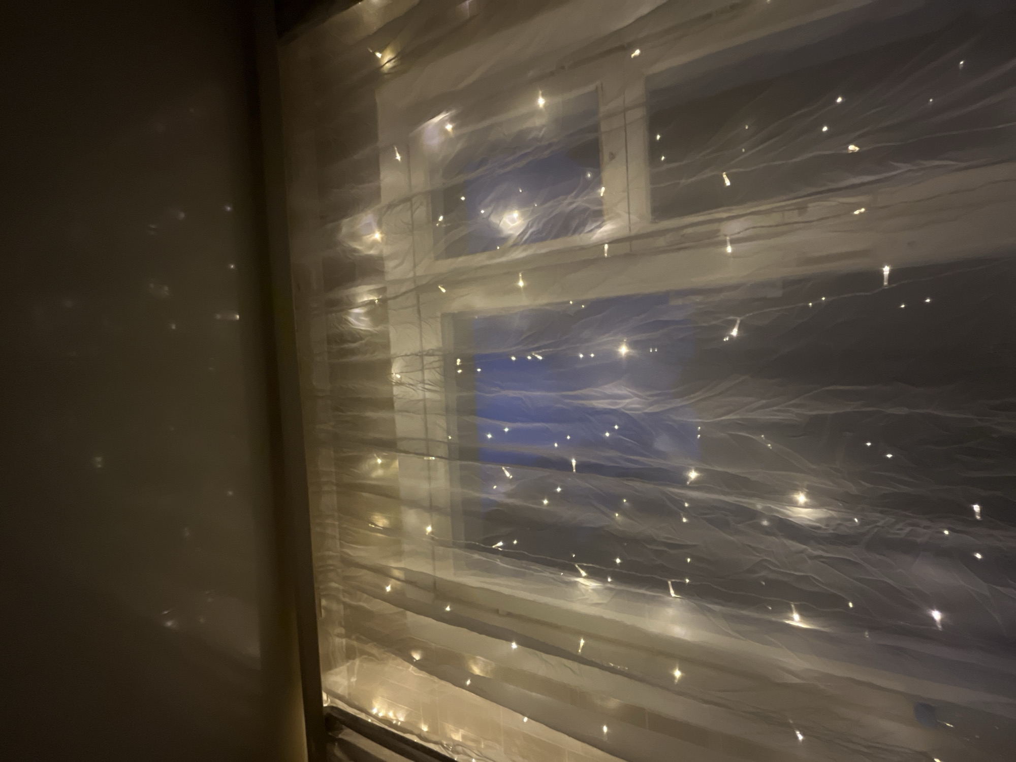 A dimly lit room in Раздолье, Russia features a window draped with a sheer, white curtain interwoven with small, warm-toned fairy lights. The window appears to be divided into multiple panes. Outside, darkness or a very low-light environment is visible. The wall to the left is a solid, neutral color, reflecting subtle light patterns. No people or actions are visible in the frame.