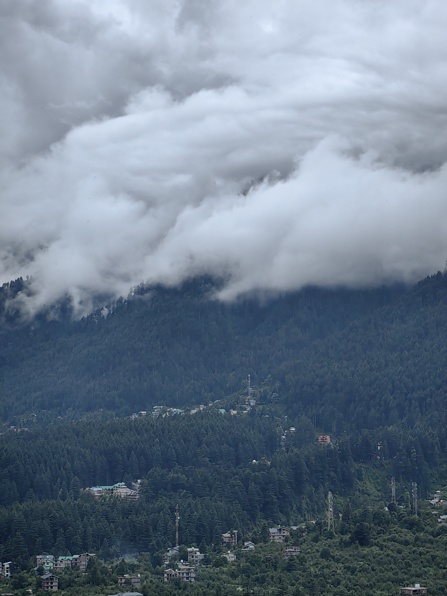 The image shows a wide shot of a mountainside densely covered with evergreen trees. The upper portion of the image is dominated by heavy, gray clouds that partially obscure the mountain peaks. Scattered throughout the forested mountainside are small clusters of buildings, suggesting a village or town nestled within the natural landscape. Several telecommunication towers are visible among the trees, indicating a populated area. The overall atmosphere is overcast and moody, typical of a cloudy or rainy day in a mountainous region. The location is identified as Vashisht, India.