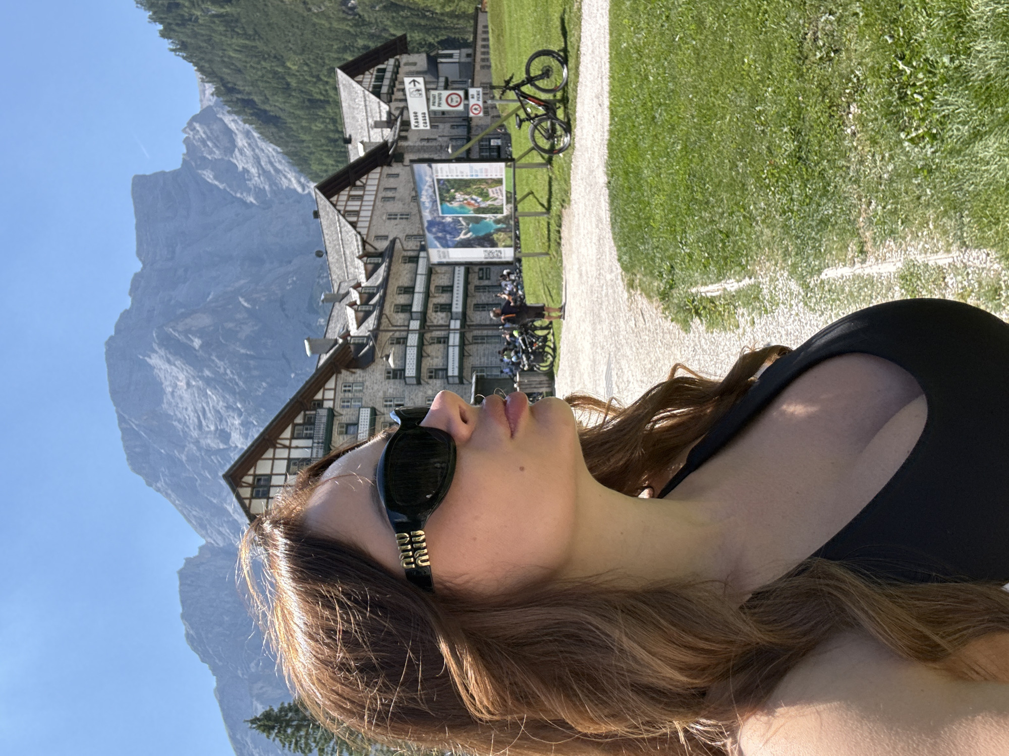 A woman with long, wavy brown hair and dark sunglasses is pictured in the foreground, facing to the left. She is wearing a black top. In the background, there are buildings with pitched roofs, a mountain range, and trees. Several bicycles are visible, and a signpost with directional arrows and text, including "Kasse" and "Privat," stands near the buildings. A man is standing in front of a large display board with images. The scene appears to be in a park-like setting with green grass and a gravel path. The sky is clear and blue. The location is identified as Prags - Braies, Italy.