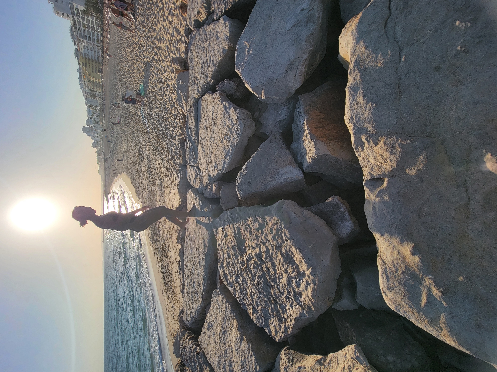 The image depicts a sunlit beach scene in Quarteira, Portugal, with the camera focused on a person standing on large rocks in the foreground. The person, a woman with her hair in a bun, is silhouetted against the bright sun and is captured mid-stride, appearing to be walking or dancing. She is wearing dark swimwear. The rocks in the foreground are rough and uneven, illuminated by the low-angle sunlight, casting shadows.
Behind the rocks, a sandy beach stretches out, dotted with people engaged in various beach activities. Further in the distance, along the coastline, a line of white buildings, likely hotels or apartments, rises against a clear blue sky. The sea is visible to the left, with gentle waves lapping at the shore. The overall atmosphere suggests a warm, sunny day at the beach, possibly during late afternoon or early morning given the sun's position.