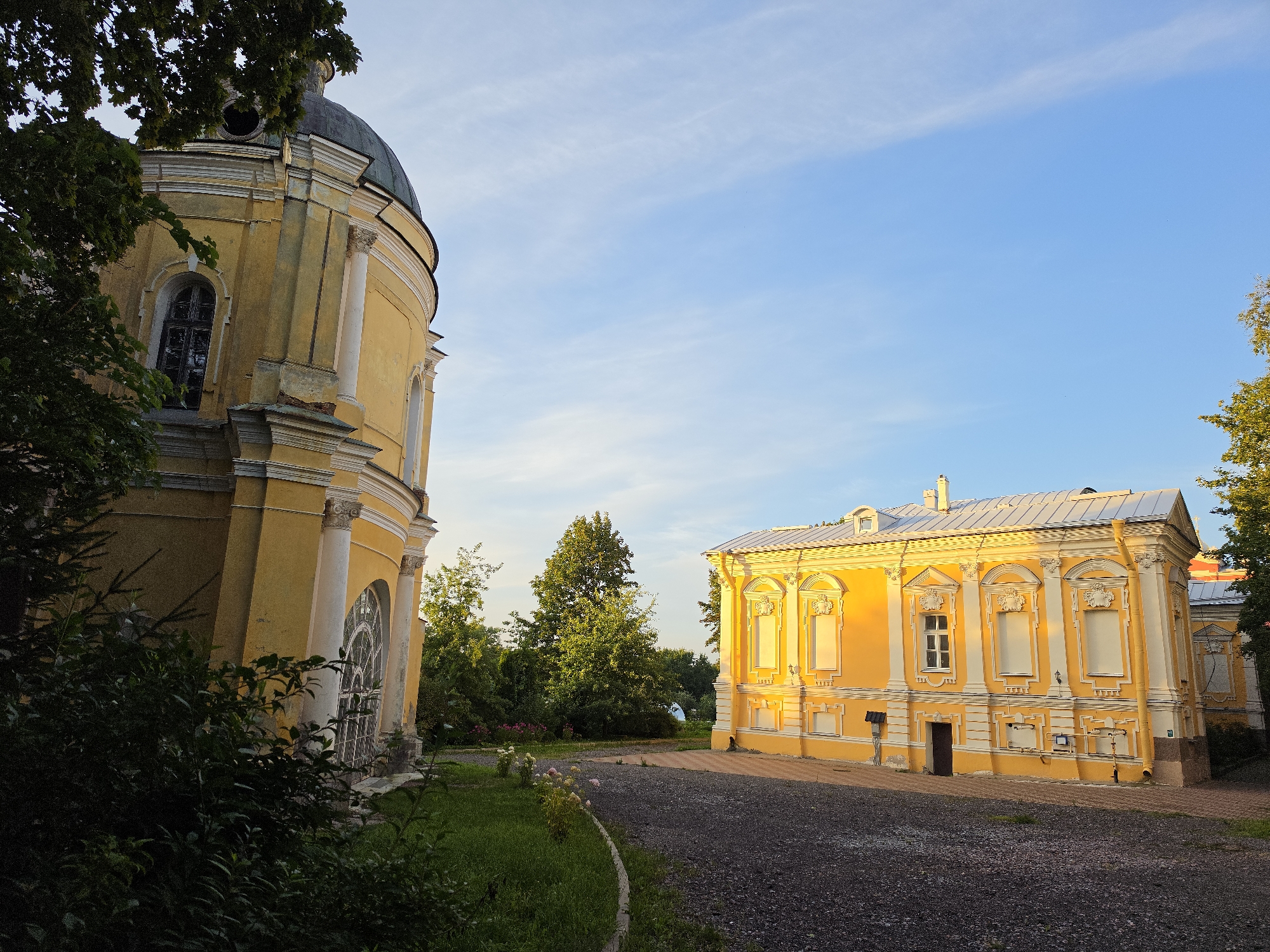 An exterior shot captures two yellow, neoclassical buildings bathed in warm, late afternoon sunlight in Strelna, Russia. On the left, a domed building with ornate white columns and large arched windows is partially obscured by dark green foliage. To the right, a rectangular building with a white roof features rows of evenly spaced windows, decorative white pilasters, and elaborate relief sculptures above each window. A gravel path leads between the buildings, with a neatly mowed lawn and a flower bed to the left of the path. Lush green trees and bushes form a backdrop. The sky above is a clear, light blue with a few wispy clouds. No people are visible in the scene.