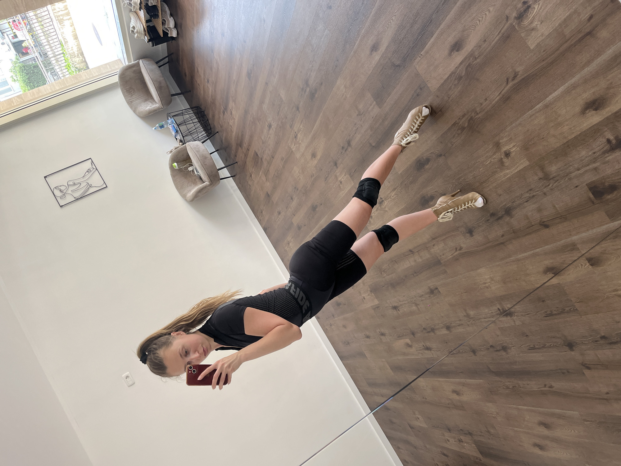 A young woman captures a mirror selfie in what appears to be a spacious, brightly lit indoor setting. She stands upright, holding a red smartphone up to photograph her reflection. Her long, light brown hair is pulled back with a black scrunchie, and she wears a black athletic short-sleeved top featuring a patterned texture on the front, with the word "REIDE" visible in grey dots. She also wears black shorts, black knee pads, and light-colored high-top sneakers with laces. Her expression is neutral as she looks towards the phone.
The room features dark brown wooden plank flooring and clean white walls. In the background, two light-colored, upholstered armchairs with black metal legs are positioned near a large window, which reveals an outdoor view of greenery and a building. Between the chairs is a black wire-frame side table holding a plastic water bottle. Another water bottle is near one of the chairs. A black metal wire art piece depicting a reclining figure adorns the white wall. A white electrical outlet is also visible on the wall. Natural daylight illuminates the scene, suggesting it is daytime. The selfie is taken in a large mirror, the bottom edge of which forms a thin, dark horizontal line across the lower part of the image.