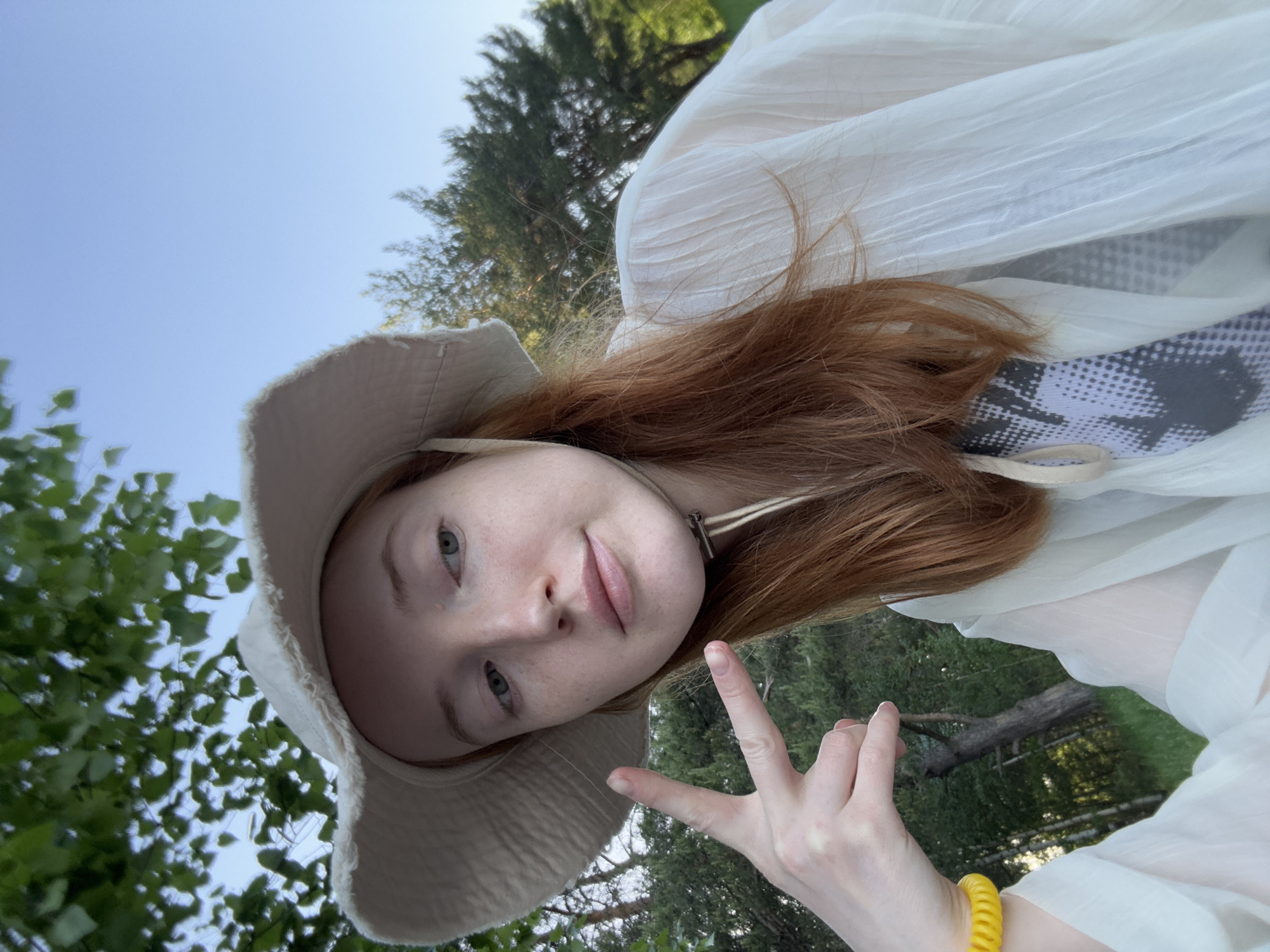 A young woman with red hair and a light complexion is taking a selfie outdoors. She is wearing a light-colored bucket hat and a white, translucent top. She is making a peace sign with her right hand. In the background, there are green trees and a blue sky.
Without precise location data, I cannot pinpoint the exact location in Chaikovsky, Russia.