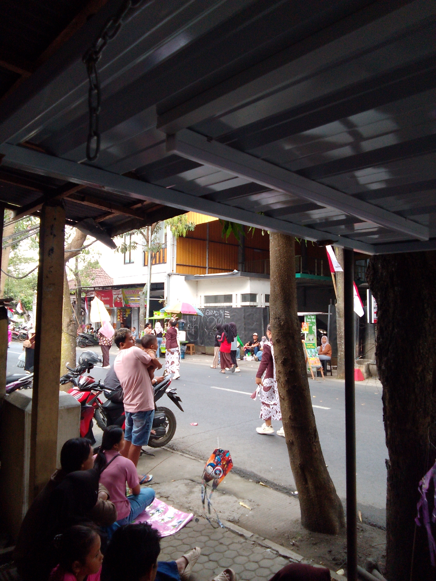 The image depicts a street scene in Trenggalek, Indonesia, possibly near the Alun-Alun (town square) or a busy marketplace, given the presence of vendors and pedestrian activity. The foreground features a covered seating area, likely a simple roadside stall or shelter, with a metal roof supported by wooden beams and a metal pole. In this shaded area, several people are seated on the ground or on a mat. A woman in a dark top and another in a pink shirt, both sitting, are visible, along with two young children. A kite with a colorful, patterned design lies on the ground nearby.
Moving towards the street, a red motorcycle is parked. A man in a light pink shirt and blue shorts stands near the motorcycle, holding a baby. He is facing away from the camera, looking towards the street. Behind him, another person is partially visible.
The street itself is moderately busy. Several people are walking and standing. A woman in a pink headscarf and patterned skirt walks on the sidewalk. Further down the street, other individuals are seen near stalls. Some are wearing traditional Indonesian attire, including headscarves and long skirts. Umbrellas are also visible, suggesting it might be a sunny day or that vendors are using them for shade. Buildings line the street, some appearing to be shops or businesses with signage. The overall atmosphere suggests a typical day in a bustling Indonesian town. The architectural style of the buildings and the clothing of the people are consistent with Indonesian street scenes.