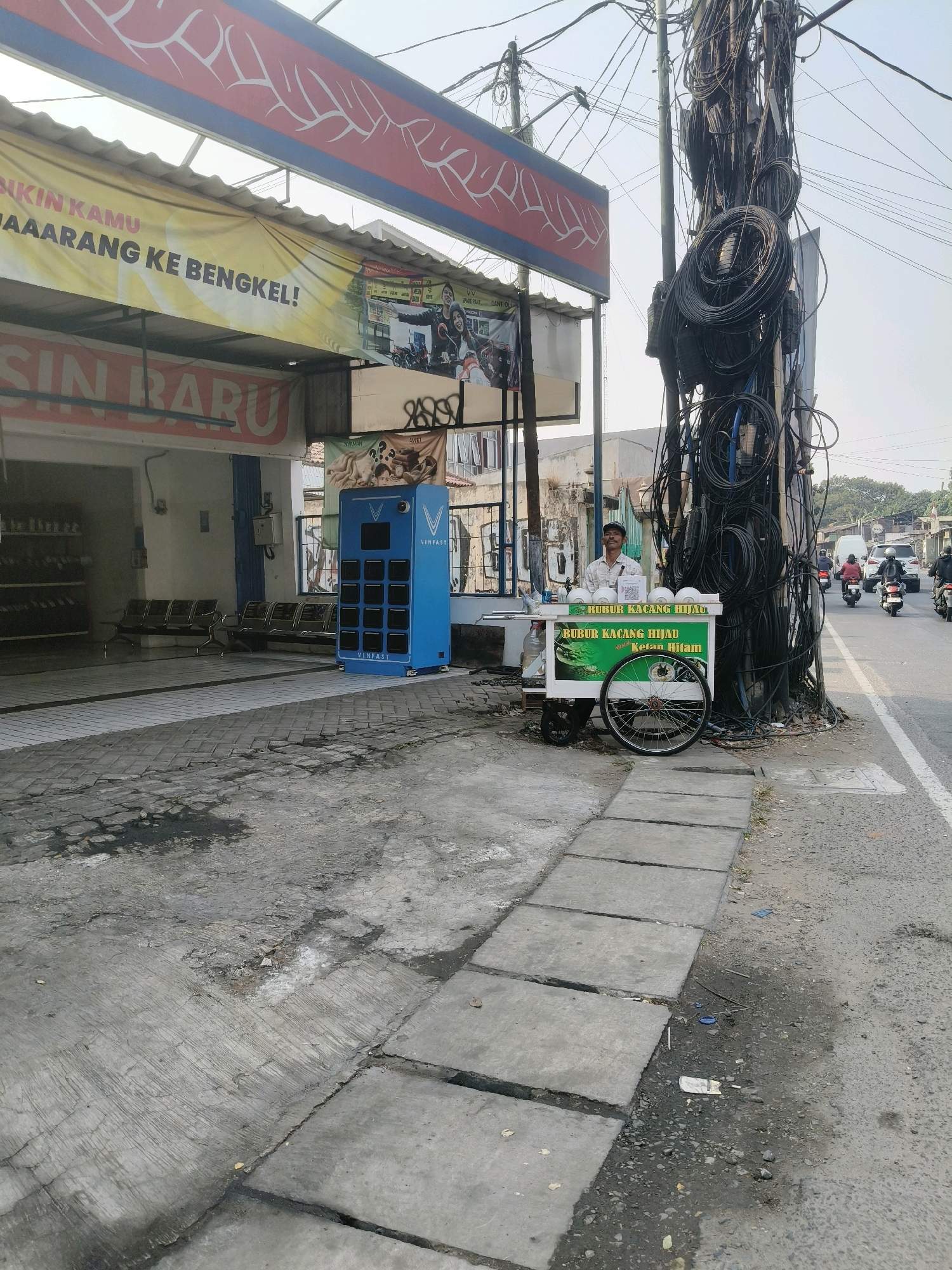 The image depicts a street scene in South Tangerang, Indonesia, characterized by a cluttered utility pole laden with electrical wires. To the left of the pole, a man stands behind a food cart selling "Bubur Kacang Hijau" (mung bean porridge) and "Bubur Ketan Hitam" (black sticky rice porridge). The cart has a green and white sign with images of the food items. Behind the food cart and the utility pole is a building with a banner advertising "SIN BARU," suggesting a new or established business. The building appears to be a workshop or service center, with seating areas visible inside. Further to the left, another banner with red and white designs is visible above. The foreground shows a cracked concrete sidewalk with some debris, leading to a paved street where several motorcycles are passing by. The overall atmosphere is that of a busy, everyday street scene in an Indonesian city.