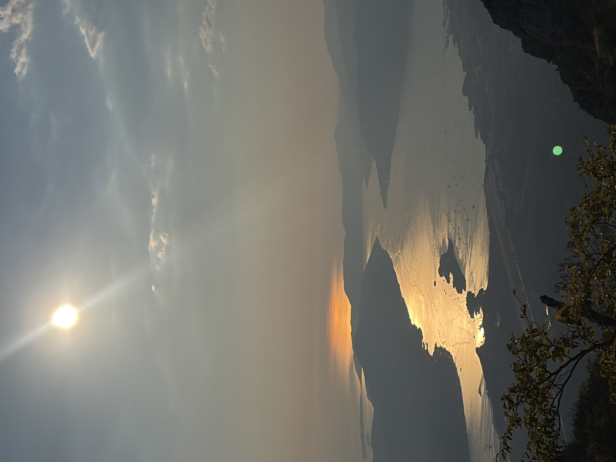 The image captures a high-angle view of a sunlit bay and surrounding mountains at sunset, presumably near Kuk, Montenegro. The sun, a bright orb, is positioned in the upper left quadrant of the sky, casting a strong reflection that shimmers across the water. The water, likely part of the Bay of Kotor, is dotted with numerous small boats, suggesting moderate maritime activity. Lush, dark green foliage occupies the lower left and right foreground, framing the vista. The distant mountains are silhouetted against the hazy, warm-toned sky, indicating a serene, natural landscape. A faint green circular object is visible in the lower right quadrant.