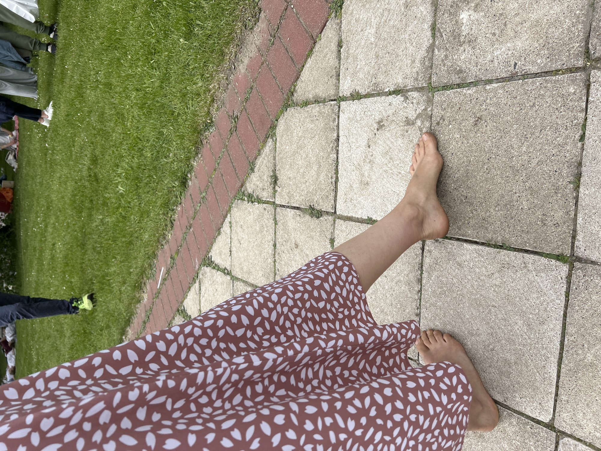 A person's legs and bare feet are visible on a gray stone tile surface. The person is wearing a brown dress with a white pattern. To the left, there's a strip of grass and a red brick border. In the background, partially visible, are other people standing on the grass.