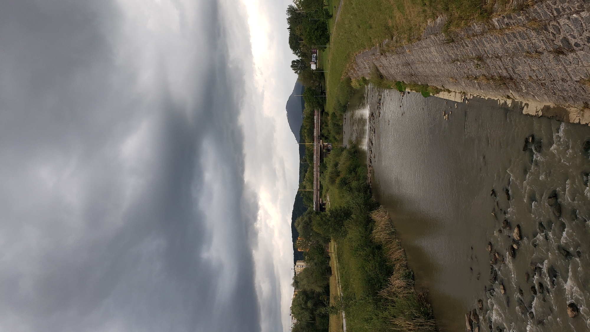 The image depicts a river flowing through a park-like area in the city of Prešov, Slovakia. The river has a rocky bed, and the water appears somewhat murky. The banks of the river are lined with green grass and dense vegetation, with a concrete embankment on the right side of the frame. In the background, a long bridge spans the river, with trees and some buildings visible beyond it. A prominent hill with a conical peak stands in the distance to the right of the bridge. The sky is overcast with dark, dramatic clouds, suggesting an impending storm. There are no people visible in the image. The scene is a natural landscape integrated into an urban environment, showcasing the confluence of nature and infrastructure. The location is identified as the Sekčov district, with the bridge likely being part of the main road connecting to the city center.