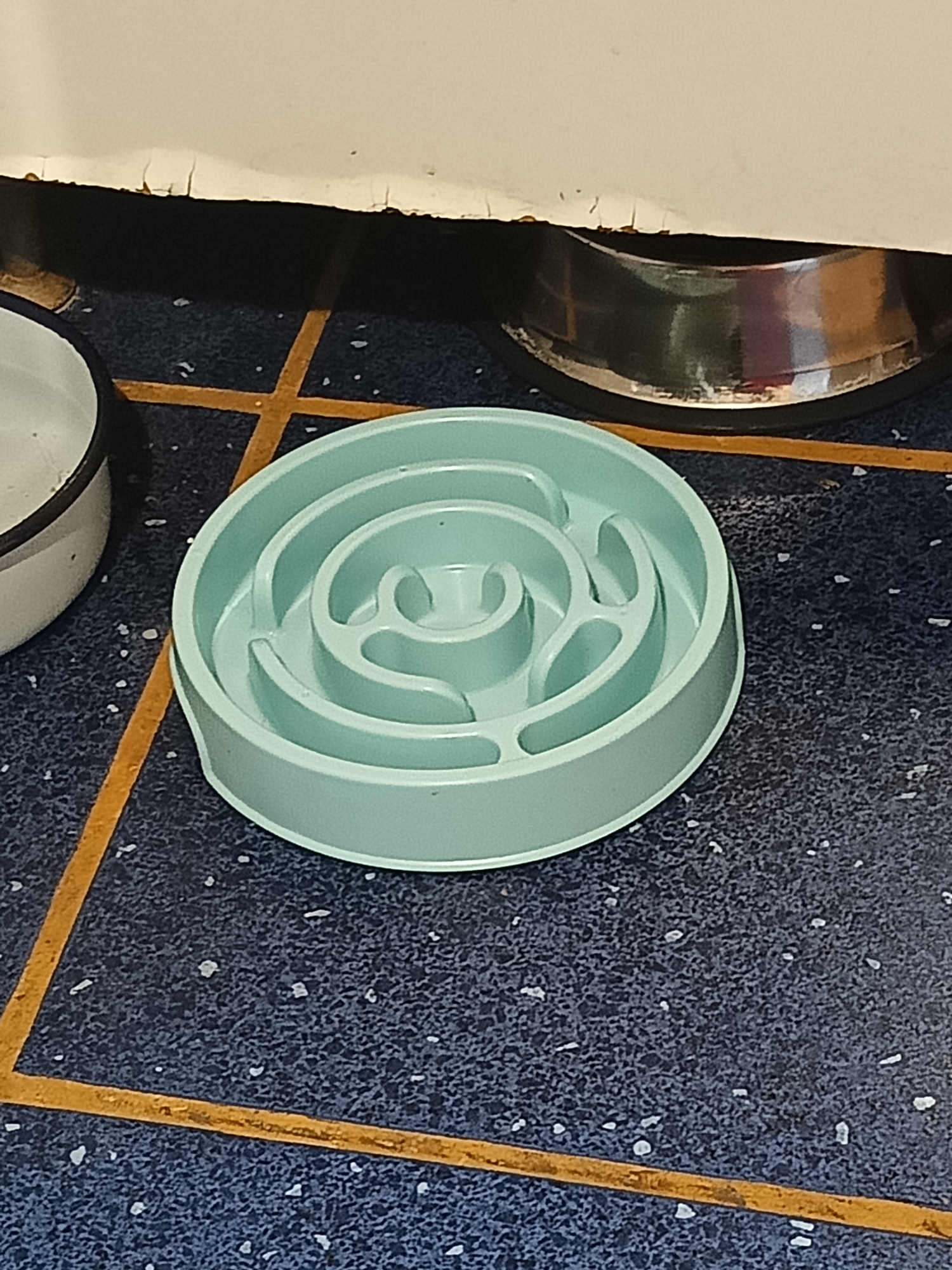 A light teal-colored dog food maze bowl is positioned on a dark blue speckled floor. The bowl features a spiral design with various compartments and raised dividers, intended to slow down a pet's eating pace. To the left of the maze bowl, a portion of a white and black pet bowl is visible. A thin, orange line runs diagonally across the floor, intersecting with another implied line to form a grid pattern. In the background, the bottom edge of a white cabinet or appliance is present, with a metallic bowl beneath it. The scene appears to be indoors, possibly in a kitchen or dining area. The location is the city of Varna, Bulgaria. There are no people or animals visible in the image.