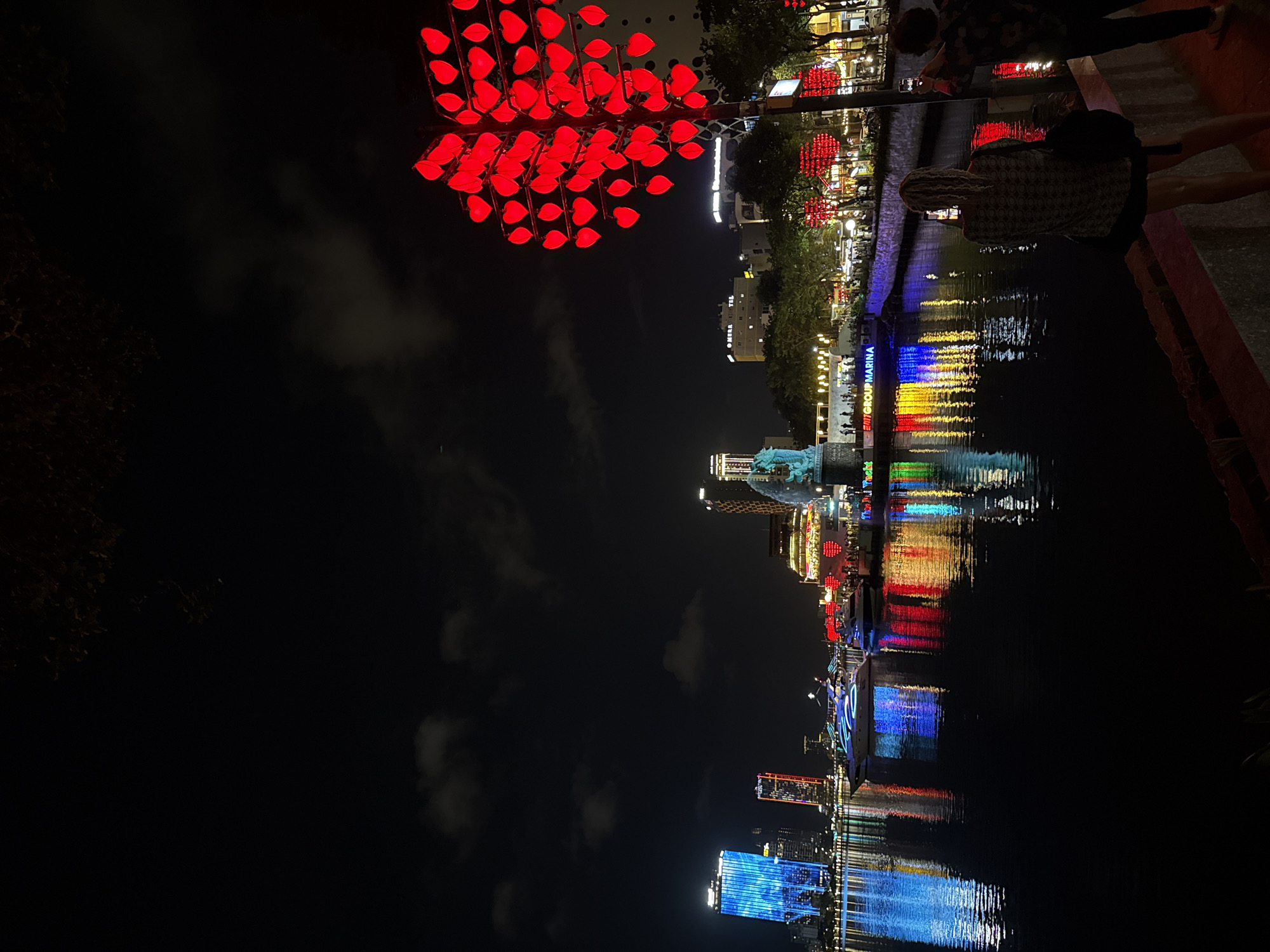 At night, the image presents a waterfront scene in Đà Nẵng, Vietnam. Dominating the upper left is a large tree-like structure adorned with red, heart-shaped lights. Adjacent to it, lights line the riverfront, where people are walking. The water reflects the city lights in vertical streaks of blue, yellow, and red. Buildings of various heights are visible in the background, illuminated against the dark sky. In the foreground, a person is standing close to the edge, near the river.