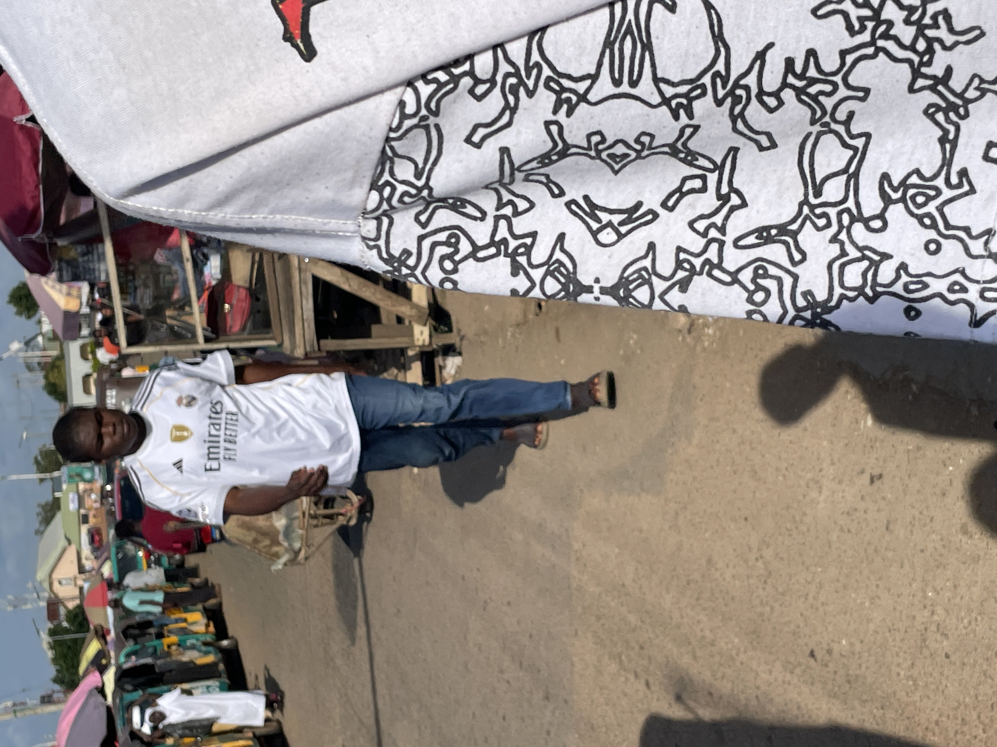 A young man walks on a paved street in Gura Top, Nigeria. He is wearing a white soccer jersey with the "Emirates Fly Better" logo and blue jeans. He is carrying a dark object. There are market stalls and vehicles in the background. The scene appears to be during the day, under a bright sky.