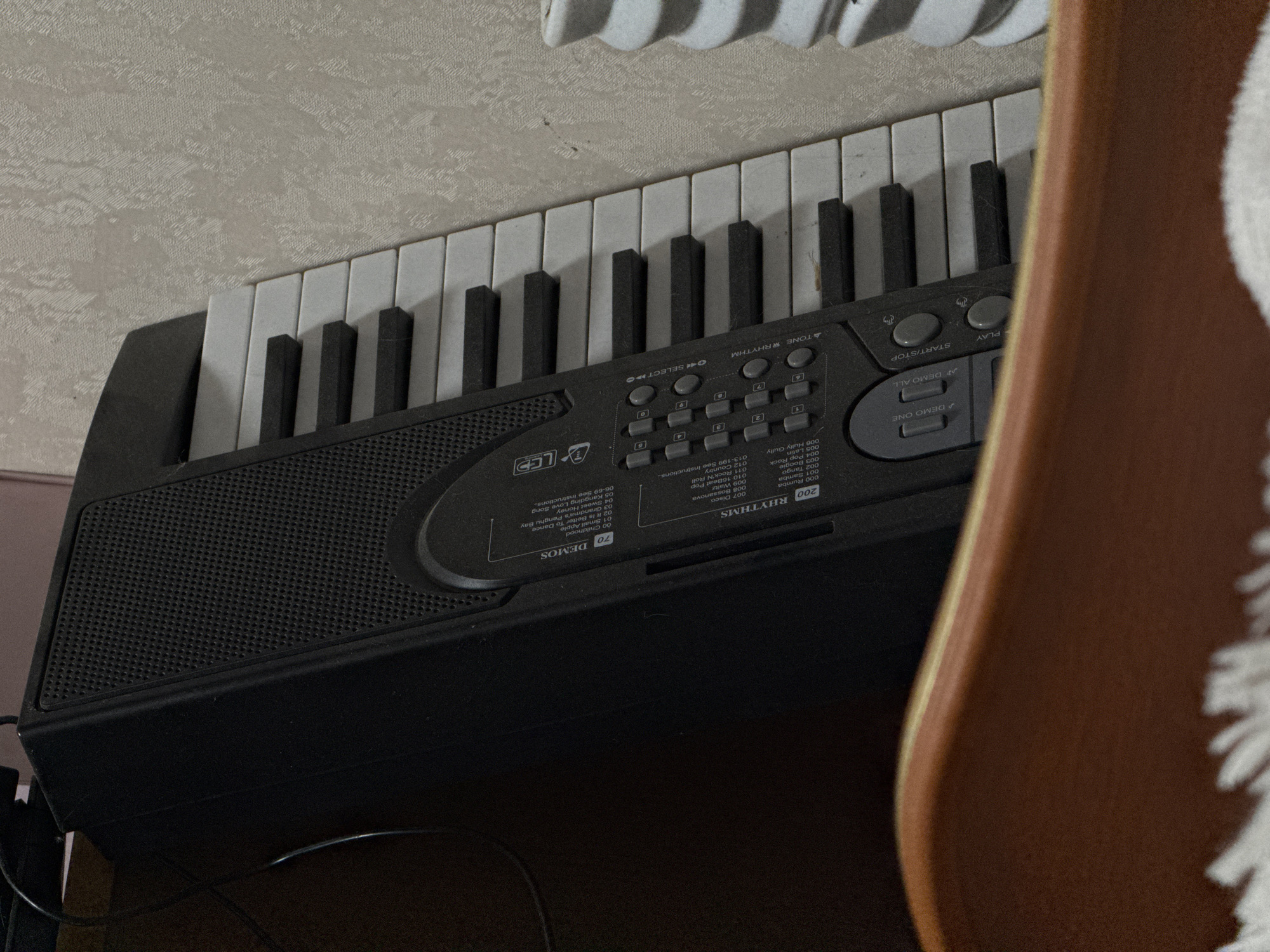 A close-up shot focuses on a black electronic keyboard with white and black keys, positioned at an angle against a light-colored wall. The keyboard's control panel displays various buttons and printed labels indicating functions such as "RHYTHMS," "DEMOS," "START/STOP," and "PLAY," alongside a numerical keypad and an LED display. Text indicating various rhythm options like "Rumba," "Samba," "Tango," "Boogie," and "Pop" is visible. A wooden chair back partially obscures the right side of the keyboard. The image provides no specific information that would indicate it was taken in Izhevsk, Russia, or any information about people or actions within the scene. The description is limited to the visible elements of the image.