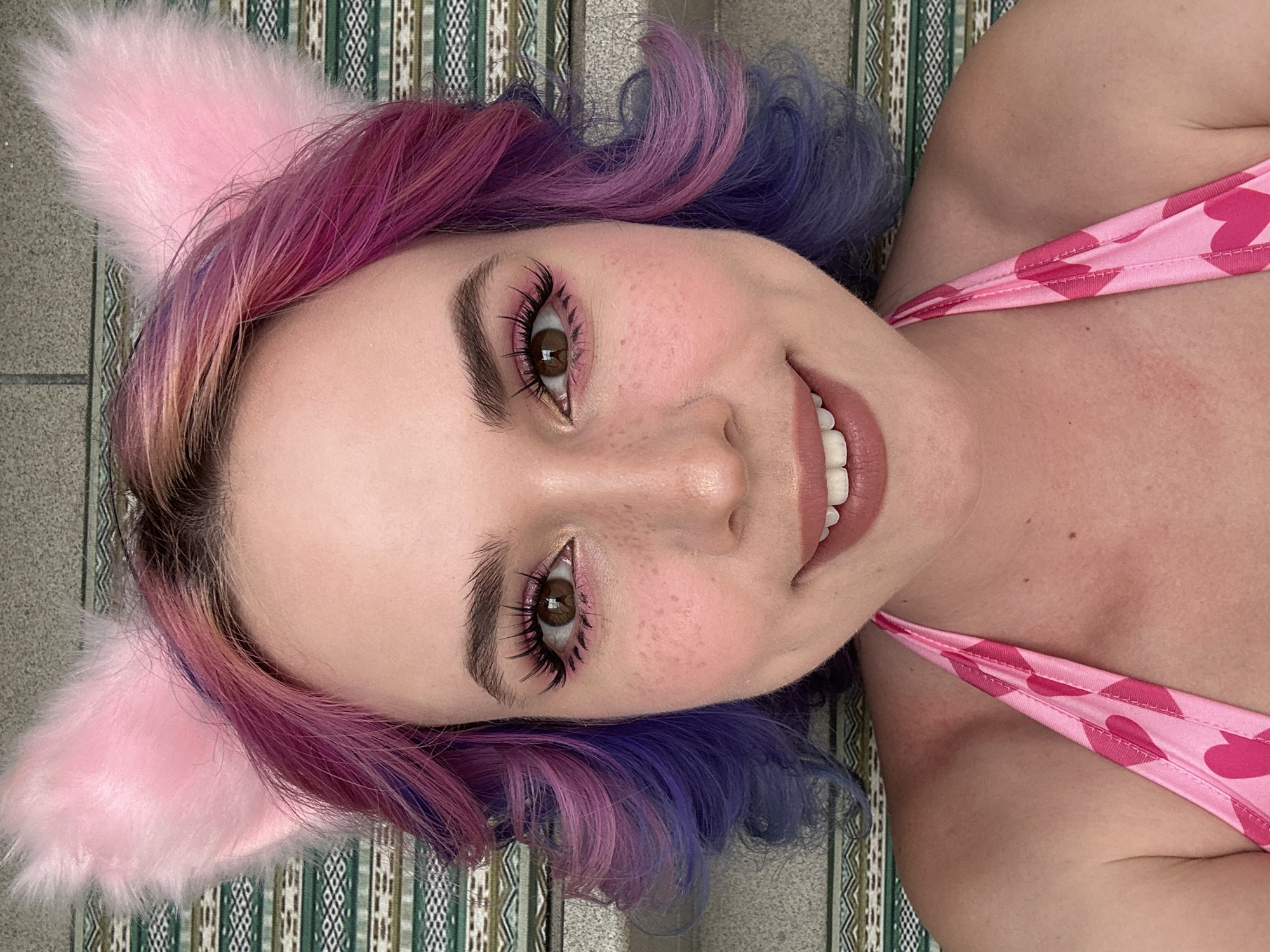 A woman with pink cat ears is lying on a striped patterned surface. She has curly hair that is dyed pink and blue. She is wearing a pink top with hearts.