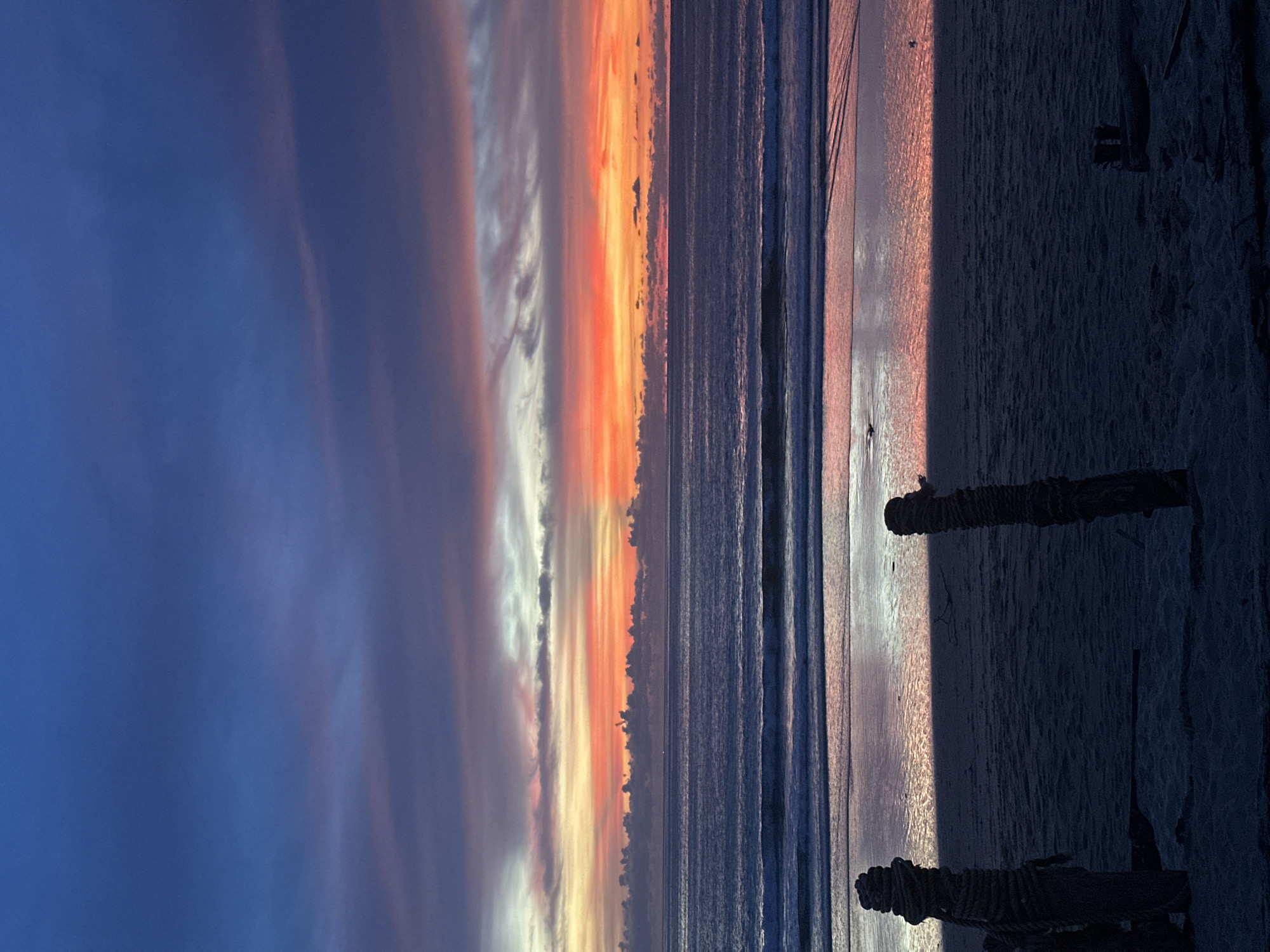 This is a vertical, medium shot of a beach at sunset. The sky is a deep blue at the top, transitioning to streaks of pink, orange, and yellow as it meets the horizon. Wispy clouds are scattered across the sky, catching the sunset colors. The ocean is a deep blue with small waves rolling towards the shore. The wet sand reflects the colors of the sky and ocean. In the foreground, two weathered wooden posts wrapped in rope stand on the beach, casting dark shadows on the sand. The scene is peaceful and serene.