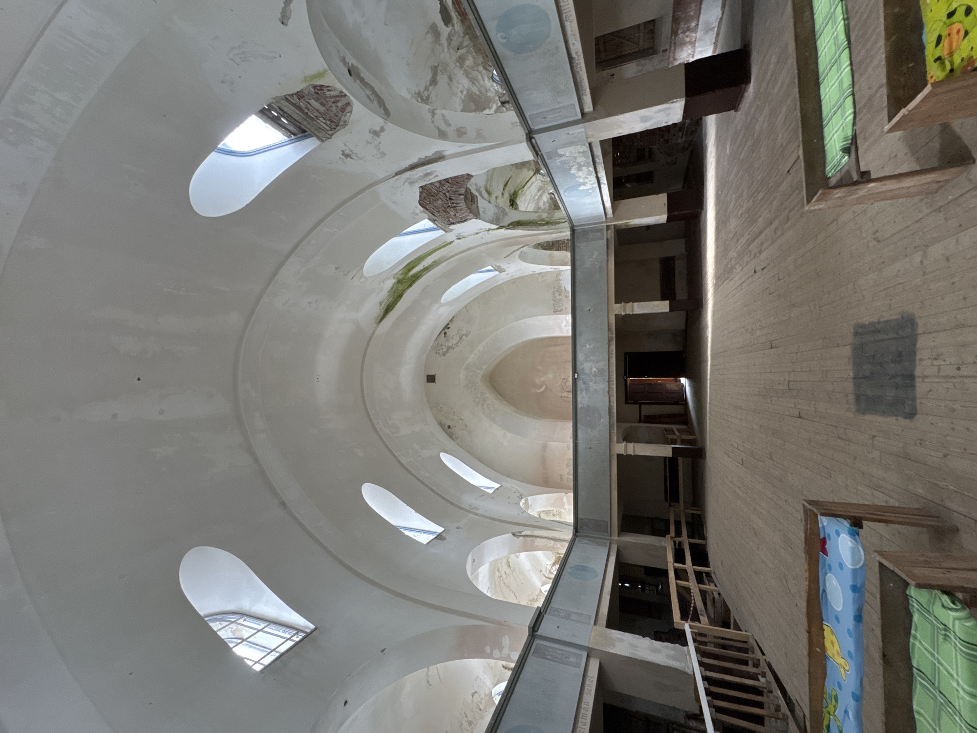 The image shows the interior of a large, dilapidated building, possibly a former place of worship or public gathering space, located in Lumivaara, Russia. The architecture features high, arched ceilings with multiple arched windows, some of which are broken, allowing natural light to enter. The walls and ceiling show signs of decay, with peeling paint, water stains, and visible brickwork in some areas.
The main floor is constructed of wooden planks and appears to be empty except for a few makeshift beds or cots at the foreground, equipped with colorful blankets. A wooden railing system is visible on the left side, leading up a ramp or stairs. A balcony or mezzanine level runs along the sides of the building, supported by pillars. The space is illuminated by the natural light filtering in from the windows, creating a somber and abandoned atmosphere. The overall impression is one of neglect and disuse. There are no people visible in the image.