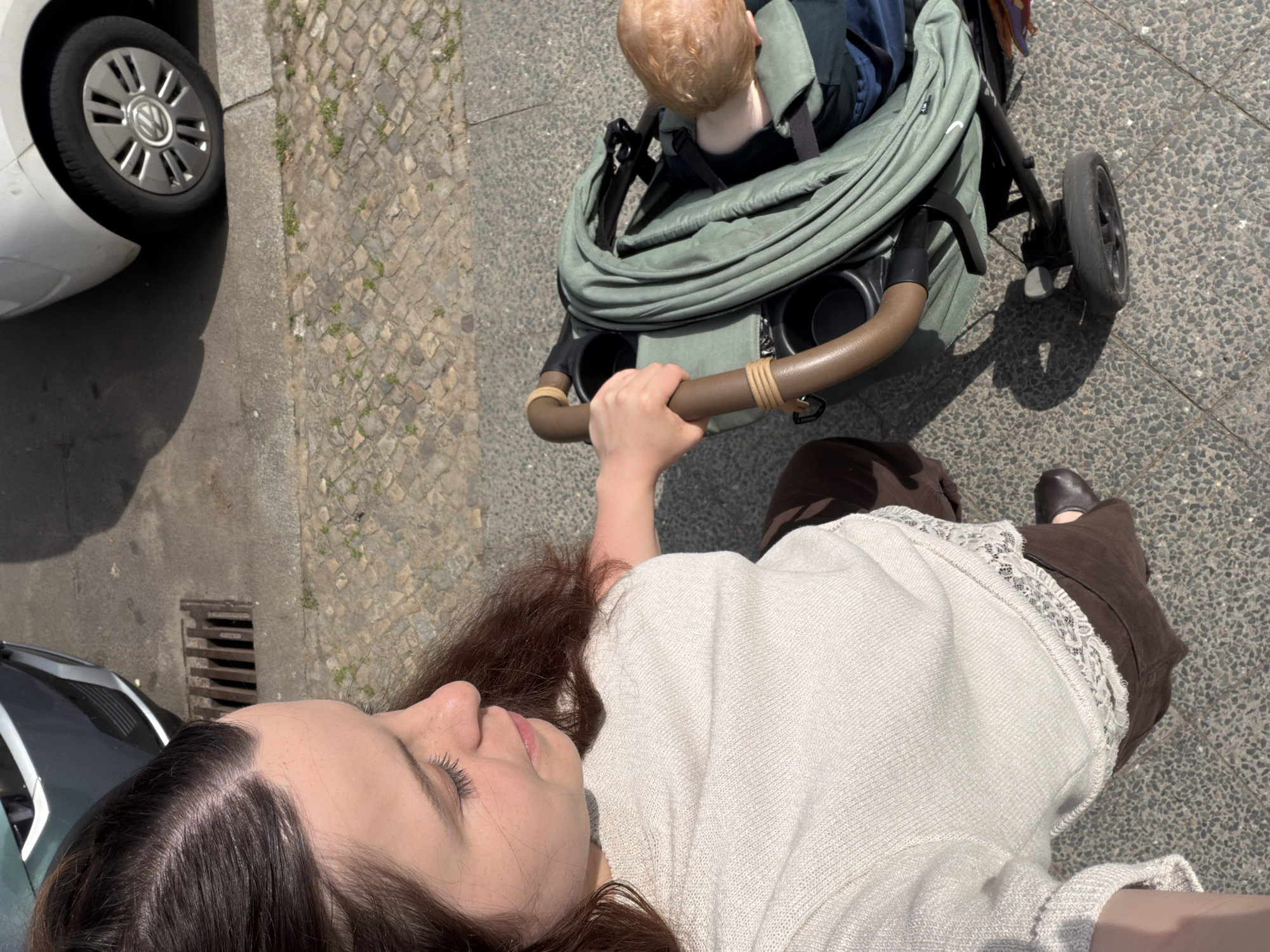 A woman pushes a baby stroller on a sidewalk. The stroller is green and has a brown handle. The baby is fair-skinned and sitting in the stroller. The woman has dark hair, a white shirt, and brown pants. There is a car parked on the left side of the image. The sidewalk is made of stone blocks. The image appears to be taken in Berlin, Germany, as suggested by the sidewalk construction style.