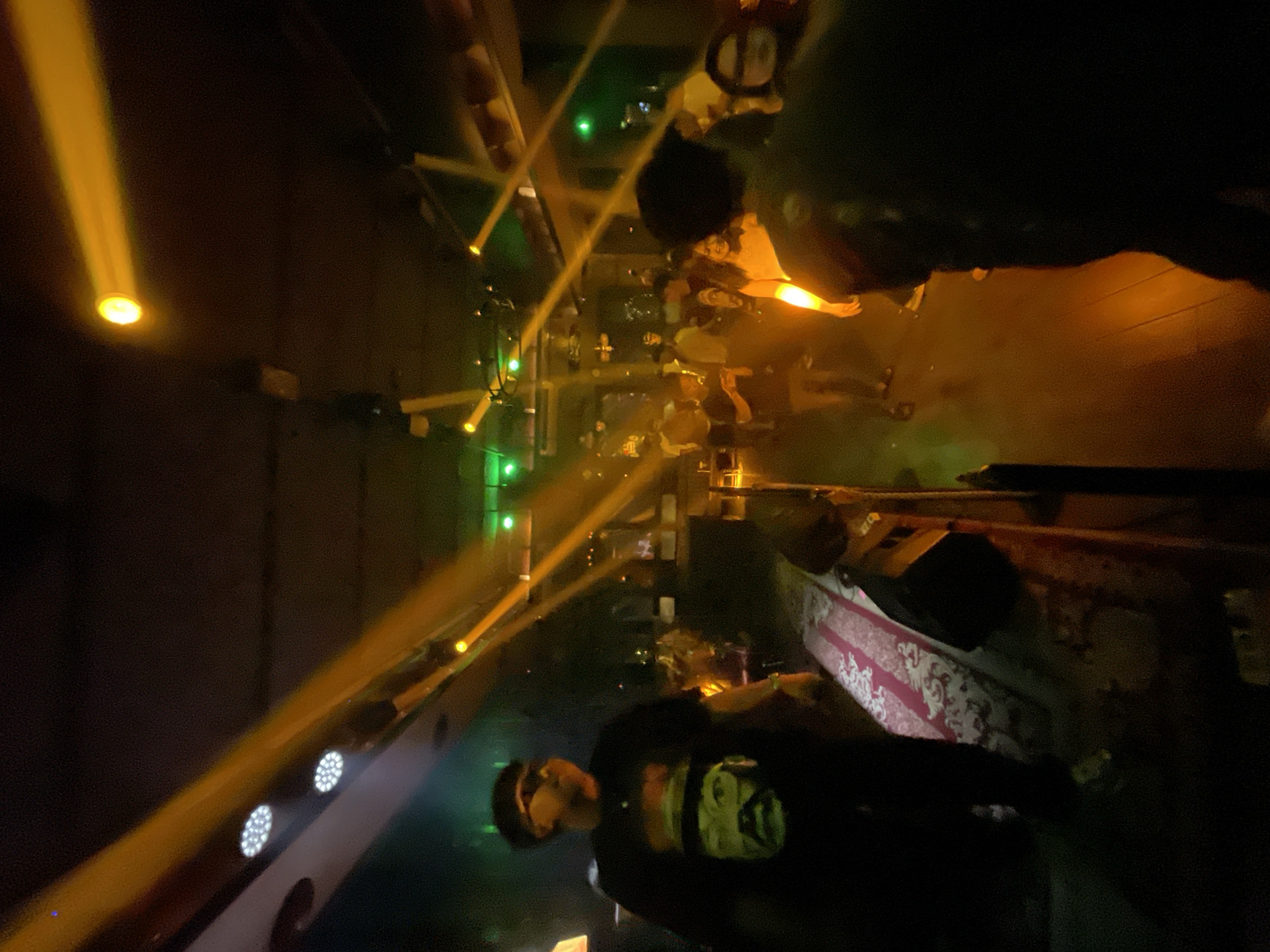 The image depicts a dimly lit indoor venue, possibly a nightclub or bar, with several beams of yellow and green light cutting through the darkness from overhead spotlights. In the foreground on the left, a person wearing sunglasses and a dark t-shirt with a graphic print of a face is visible. Further into the scene, other individuals are present, some standing and some appearing to move. The floor appears to be made of wood, and there is a red and white patterned rug laid out in the middle ground. The overall ambiance suggests a social gathering or event taking place in Central Jakarta, Indonesia.