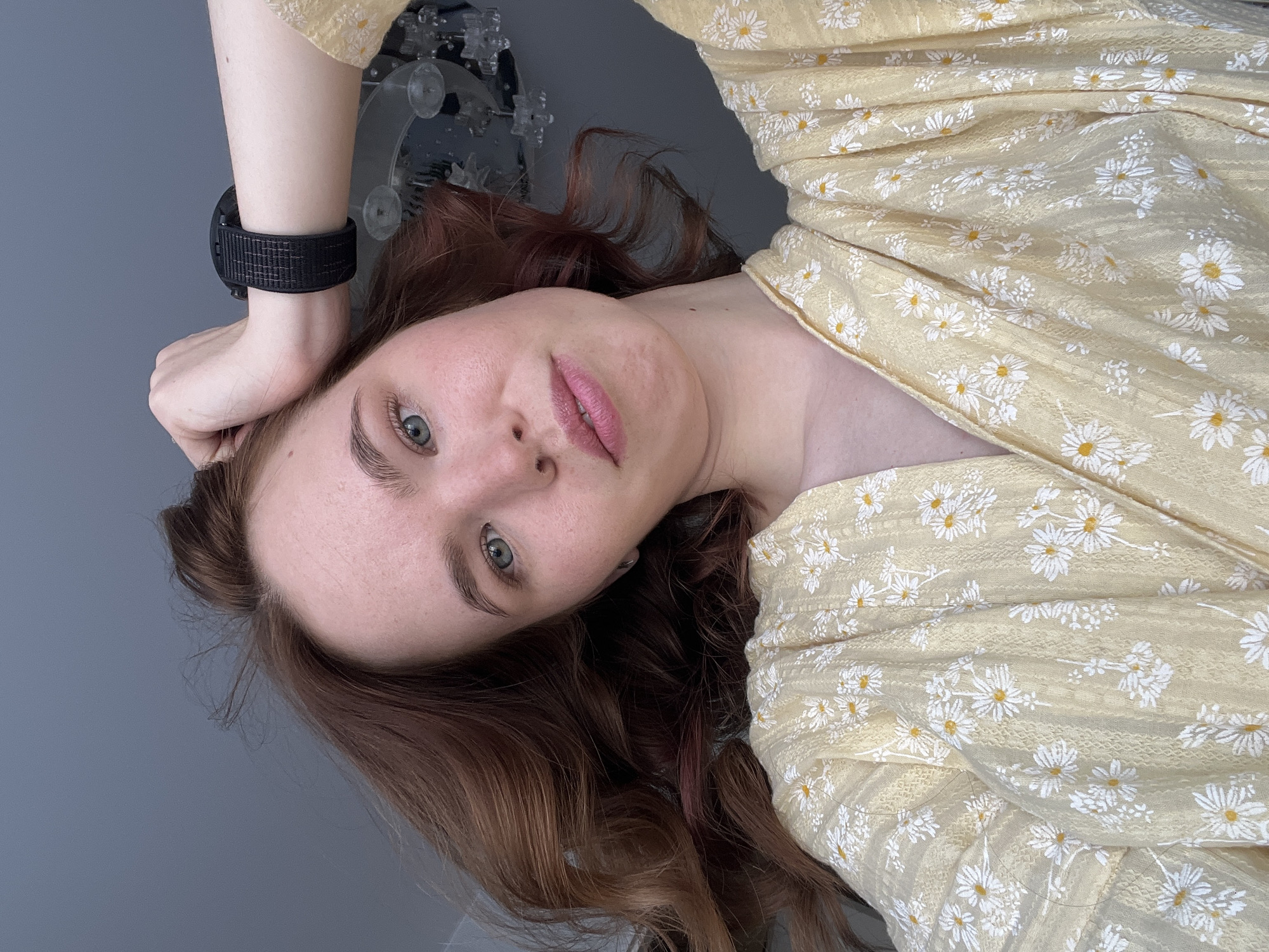 A woman with reddish-brown, wavy hair and greenish-blue eyes is depicted in a close-up, angled portrait. She wears a light yellow garment adorned with a repeating pattern of white daisy-like flowers with yellow centers. Her left hand, featuring a black digital watch with a woven strap on her wrist, is positioned near the upper left side of her head. Her lips are slightly parted, and her gaze is directed towards the viewer. The background consists of a solid light grey surface, likely a wall or ceiling, with a portion of a modern chandelier featuring multiple transparent, spherical or teardrop-shaped elements visible to the upper right. The scene takes place indoors. The specific location within the city of Chaikovsky, Russia, cannot be determined from the visual information present in the image.