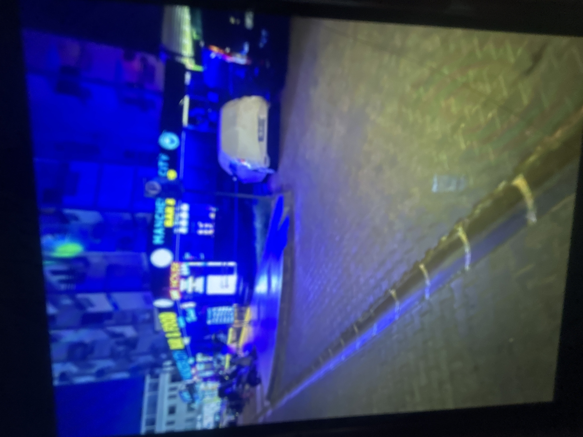 The image depicts a nighttime scene in an urban setting, likely a city street. The perspective is rotated, making it appear skewed. The left side shows illuminated buildings with various signs, some of which read "MANCHE" and "CITY." The lighting is primarily blue. A delivery bag sits on the sidewalk. The right side of the image displays a paved walkway or road with a guardrail. The overall environment suggests a bustling commercial district at night.