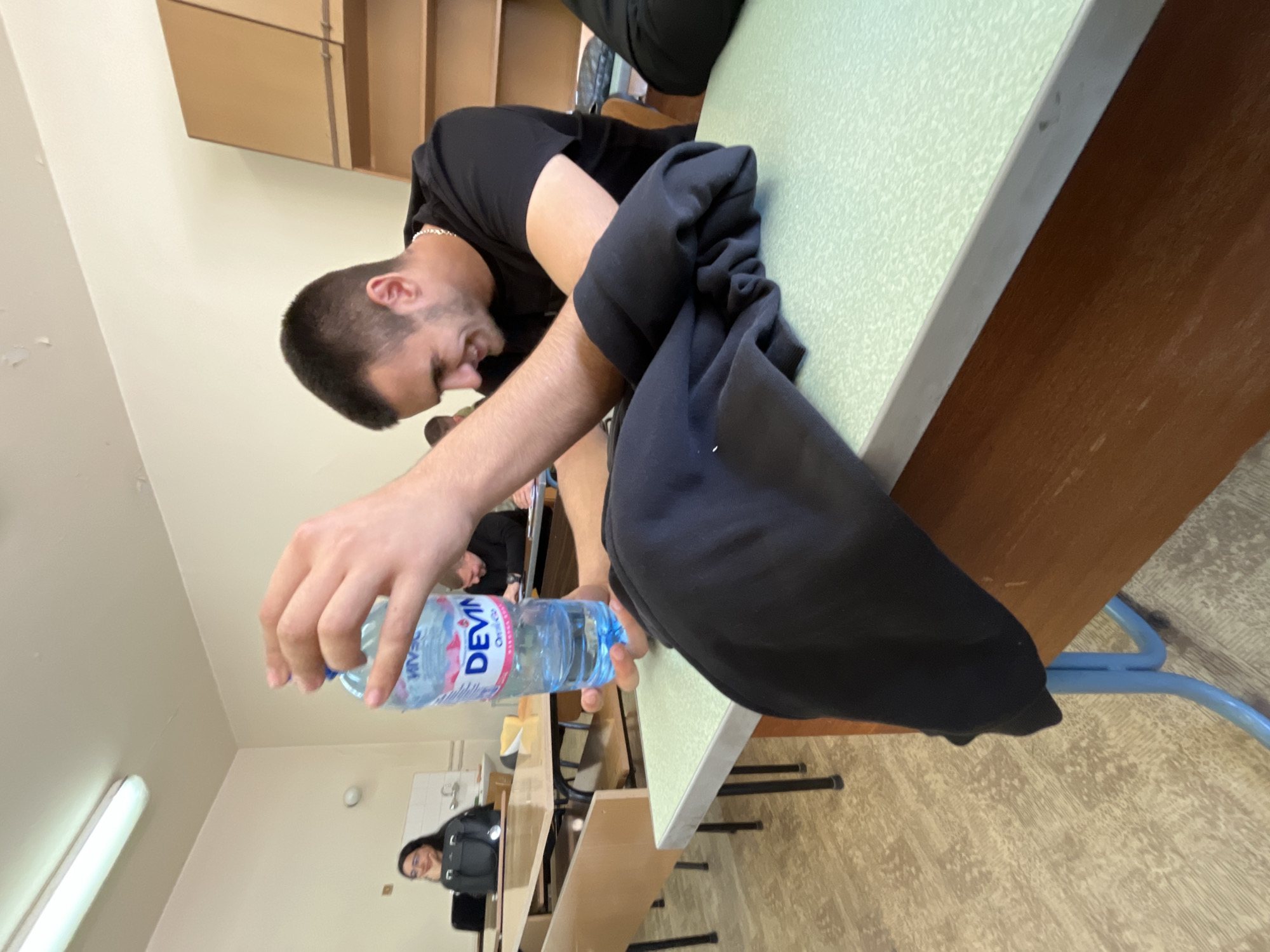 The image shows a man sitting at a desk, holding a bottle of "Devin" brand water. He is wearing a black t-shirt and appears to be in a classroom or lecture hall, possibly in Plovdiv, Bulgaria, given the brand of water. The room has other students present, desks, and institutional-style cabinets on the wall. The lighting is typical of a classroom setting. A black hoodie is placed on the desk next to him.