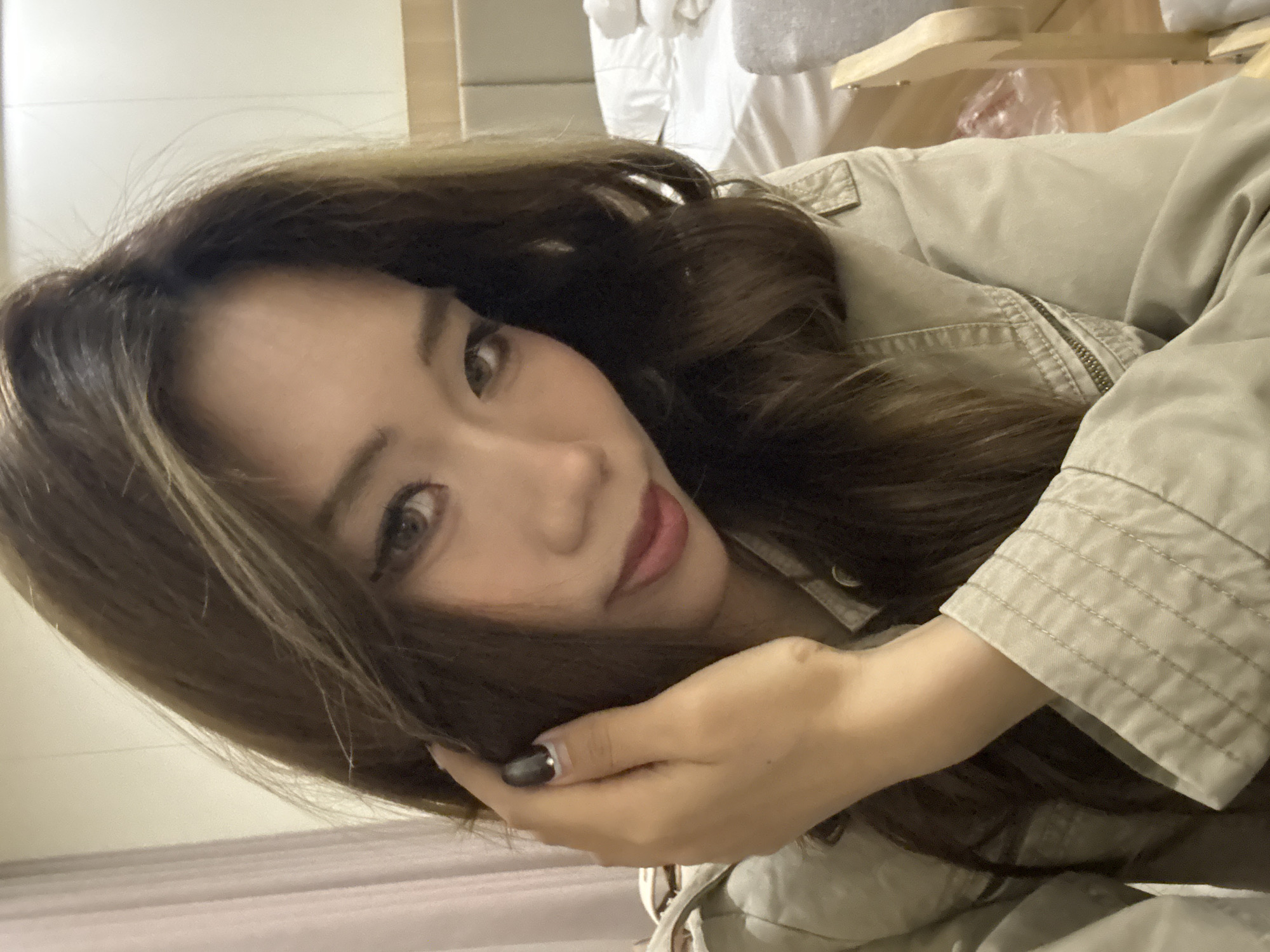 A woman with long brown hair poses closely with her hand touching her cheek. She is wearing a light brown jacket. The background appears to be an interior space with a bed and furniture. There's no specific indication that the location is Dunhuang City, China.