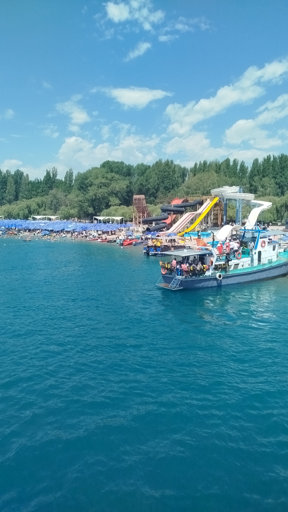 The image captures a bustling scene at a lakeside amusement park on a sunny day. In the foreground, a blue and white boat with a canopy is docked and filled with people wearing life vests. The boat has stairs leading down to the water. The lake itself is a vibrant blue, with gentle ripples on the surface.
Along the shore, a beach area is crowded with people relaxing under numerous blue umbrellas. Behind the beach, a variety of water slides are visible, including a tall, winding red and black slide, a yellow slide, and a white slide that appears to be a tube slide. The slides are constructed on a metal framework, with a brown wooden structure also present near the slides.
Beyond the amusement park structures, a dense line of green trees forms a backdrop. The sky above is a clear blue with scattered white clouds. The overall impression is of a lively recreational area filled with visitors enjoying the water and the attractions.