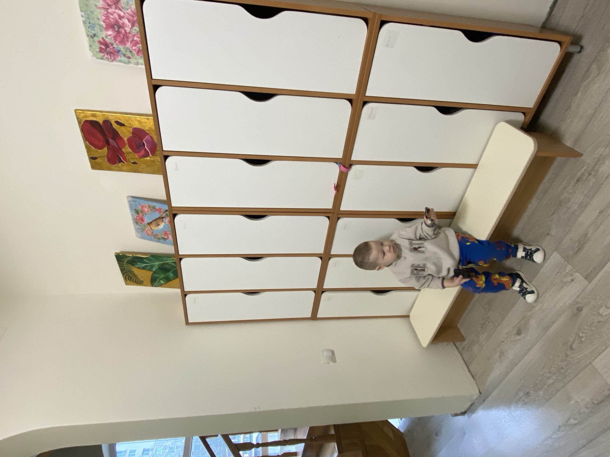 Here is a detailed description of the image:
**Scene:** The image appears to be taken indoors, possibly in a children's room or a daycare center. The setting is simple, with focus on a child and furniture.
**People:** A young boy, estimated to be around 2-4 years old, is the primary subject. He is dressed in a grey sweatshirt with the word "GREAT" in blue letters, blue pants with a cartoonish pattern, and sneakers.
**Objects:** The dominant feature is a large, multi-tiered cabinet. Each tier appears to be a storage unit or drawer. Several colorful artworks hang on the wall next to the cabinet. A bench-like seat is placed on the right side, next to the cabinet.
**Actions and Interactions:** The boy is sitting on the bench. He is holding a dark-colored object in his hands.
**Environment:** The walls are white, and the floor is covered with light-colored wood-look planks. Light enters from a source not visible in the frame.
**Location:** There is no evidence to suggest that this photograph was taken in Sochi, Russia. The details about people, objects, actions and interactions are all that can be described given the image data.