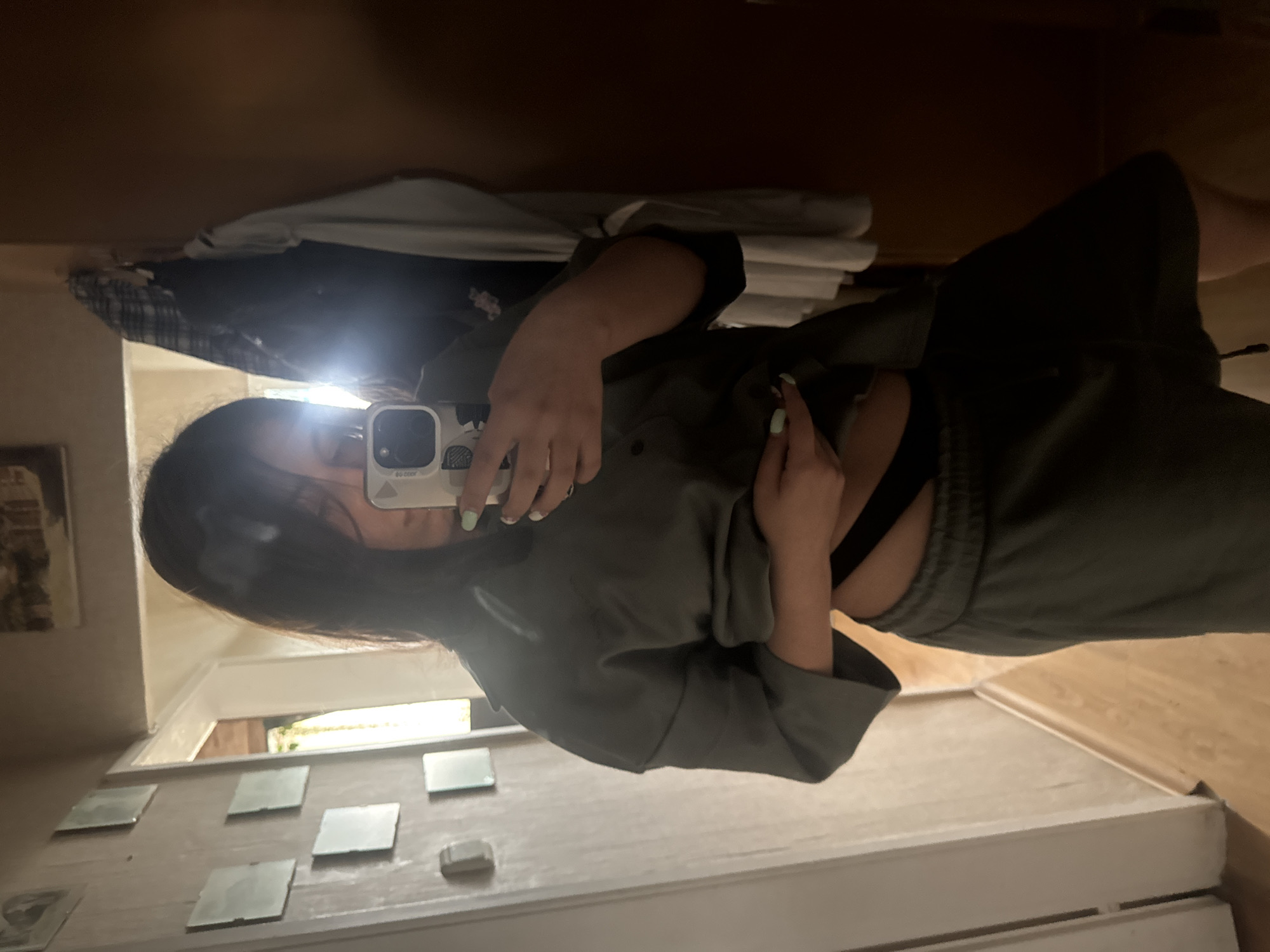 A mirror selfie, oriented clockwise by 90 degrees, captures a person standing indoors. The individual, likely female, has long, dark hair and is dressed in a dark grey or green long-sleeved top, possibly a hoodie, revealing a black sports bra underneath. She also wears matching sweatpants. One hand, adorned with light green nail polish, holds a white smartphone, its active flash creating a bright glare that partially obscures her face. Her other hand rests near her midriff.
The reflection in the mirror reveals the surrounding indoor environment. A framed picture hangs on the wall behind the person's head. To her left, various clothes, including a plaid item, are visible hanging. Natural light streams through a window, suggesting it is daytime. Below the window, several small square reflective tiles are arranged on a surface. To her right, a white object resembling a mattress or bed base is seen next to wooden flooring. Dark brown furniture or wall panels also appear in the background. Faint text, possibly "DU Osor," is partially visible on the phone case or screen. The setting is in Sofia, Bulgaria.