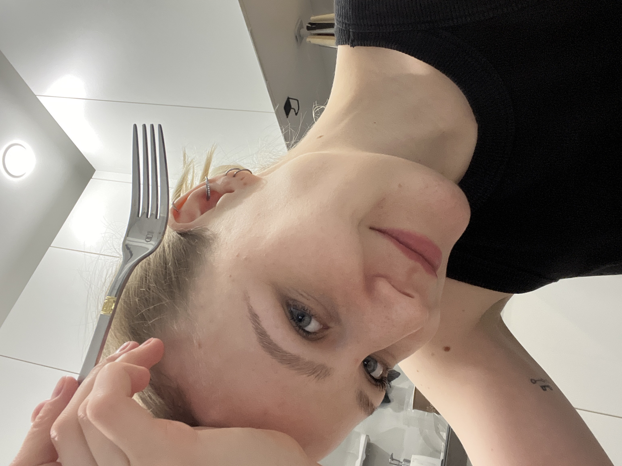 A selfie image depicts a person with light skin and blonde hair, holding a silver fork near their temple. The person wears a black tank top. Visible on their left arm is a tattoo consisting of small symbols. The background includes a white wall with a circular light fixture, suggesting an indoor setting, possibly a bathroom or kitchen. No elements in the image indicate a location in Khimki, Russia.
