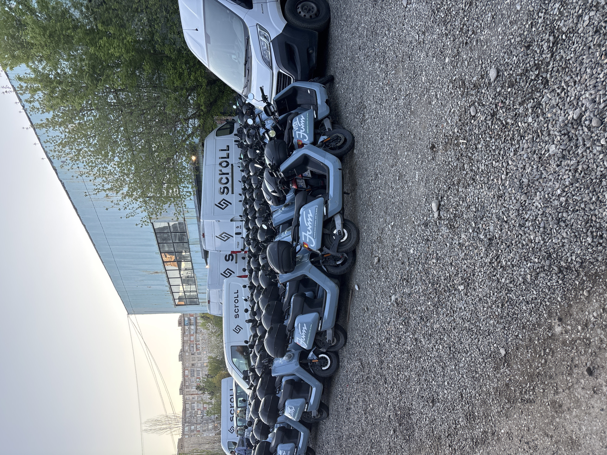 The image shows a parking area with a fleet of light blue electric scooters lined up next to several white vans bearing the logo "SCROLL." The surface is covered in gravel. A building with a metallic facade is visible in the background, along with trees and a multi-story residential building in the distance. The scene is likely located in Tbilisi, Georgia, based on the style of the residential building in the background and the presence of the "SCROLL" branded vehicles, which provide a scooter rental service in Tbilisi. No people are visible. The scooters are parked close together, suggesting they are stored or ready for rental.
