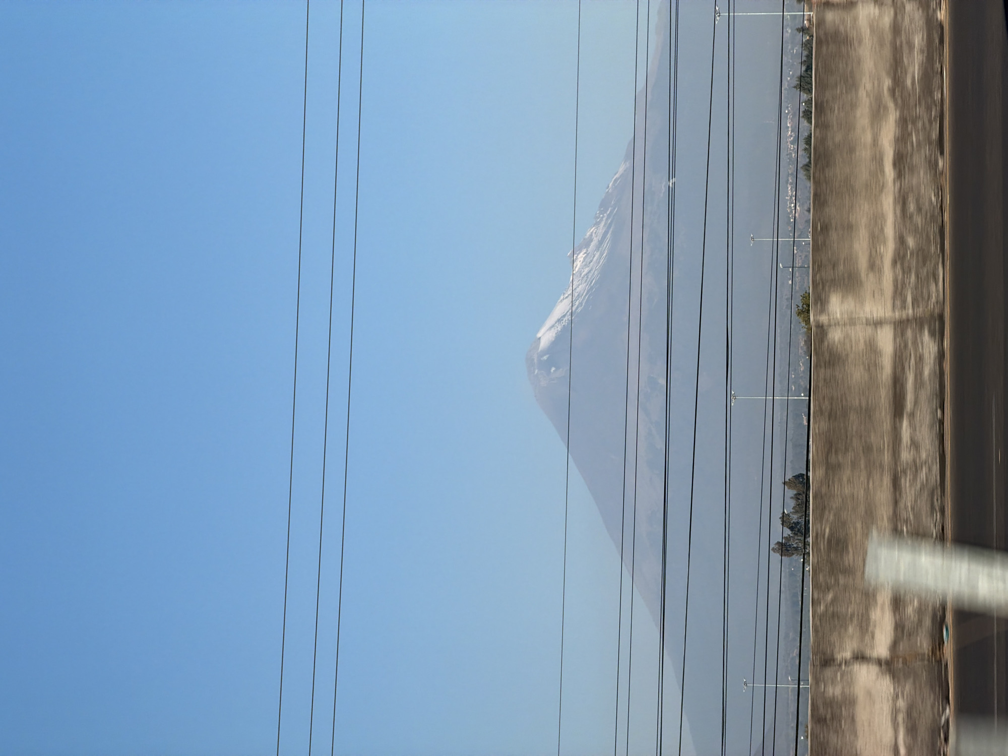 The image presents an outdoor scene likely observed from a moving vehicle in or near Santa María Zacatepec, Mexico. The central background is dominated by a large, snow-capped stratovolcano (Popocatépetl), its peak covered in white snow against a clear, light blue sky. Atmospheric haze partially obscures the mountain's lower slopes and the distant landscape. A horizontal band in the mid-ground reveals distant urban or suburban development, characterized by scattered structures and green tree cover. Several tall, slender streetlights or utility poles are visible in this mid-ground area. The immediate foreground features a concrete barrier, weathered and textured, running horizontally across the lower portion of the frame. Below this barrier, a dark road surface is visible, blurred due to motion. Multiple dark, horizontal utility lines traverse the entire image width, layered over the sky, mountain, and mid-ground elements. A single, light-colored, vertically oriented object, heavily blurred, is partially visible in the lower-left foreground, consistent with an object passed at speed. No individuals are depicted.